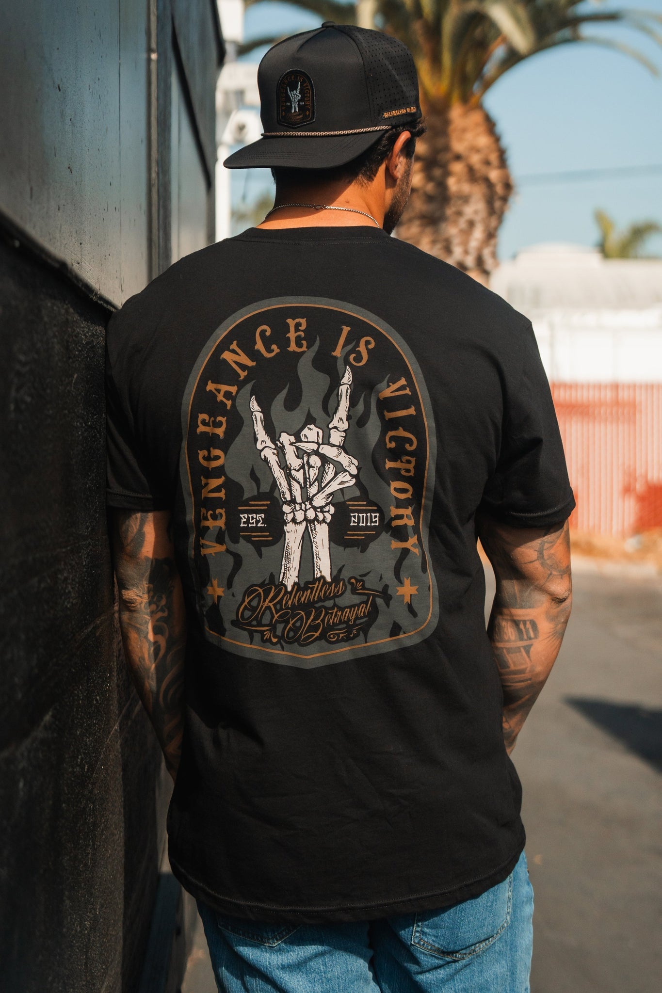 A man outdoors wears a black cap and the Vengeance Tee from Relentless Betrayal, featuring a graphic of a skeletal hand holding a torch. The scene of palm trees and distant buildings symbolizes his journey of resilience and determination, embodying the shirts theme: Vengeance is Victory.