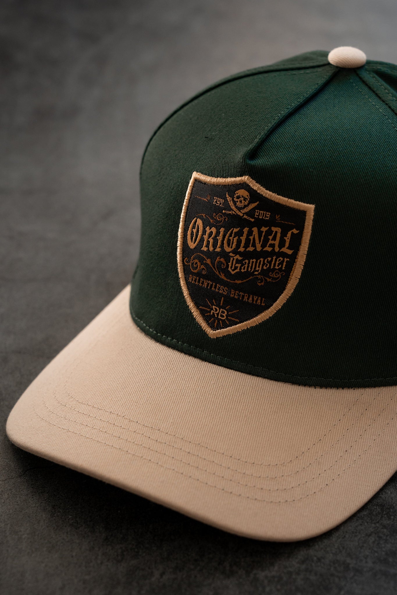 The OG Forest Green Snapback from Relentless Betrayal features a curved brim and a stylish Original Gangster patch on the front, complemented by subtle lettering and graphics. This forrest green and beige cap exudes effortless style while resting on a dark surface.
