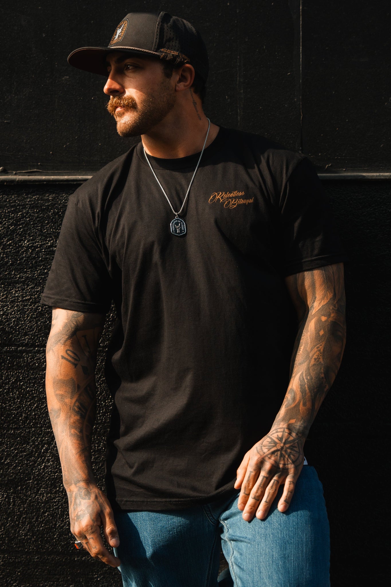 A tattooed man leans against a black wall, exuding resilience and determination in a Vengeance Tee by Relentless Betrayal, paired with jeans, a black cap, and a necklace.