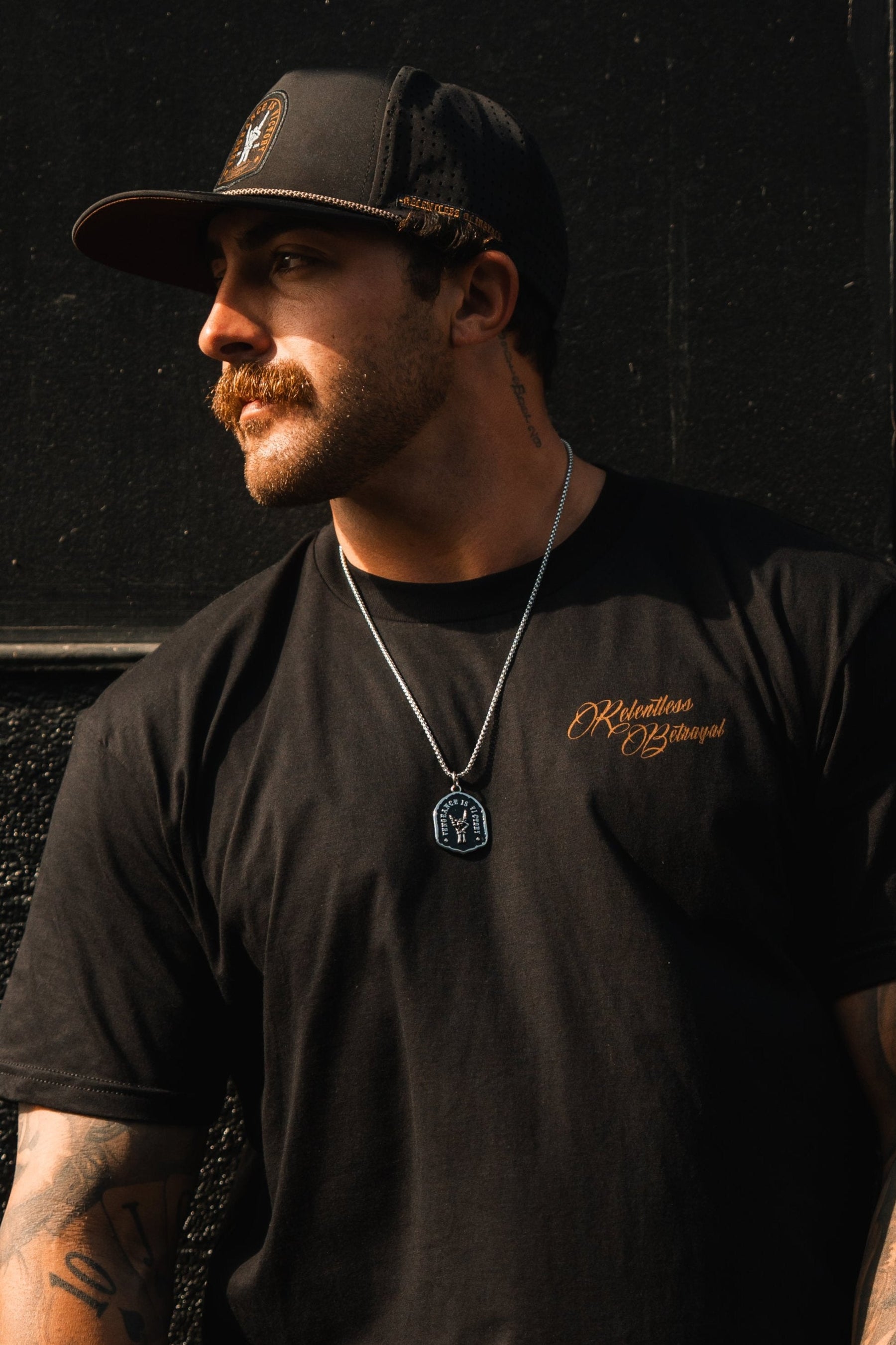 A tattooed man with a mustache, wearing a black cap and t-shirt, leans against a dark wall. The t-shirt proclaims Relentless Strength, resonating resilience. Around his neck is the Vengeance Necklace by Relentless Betrayal, completing his bold look.