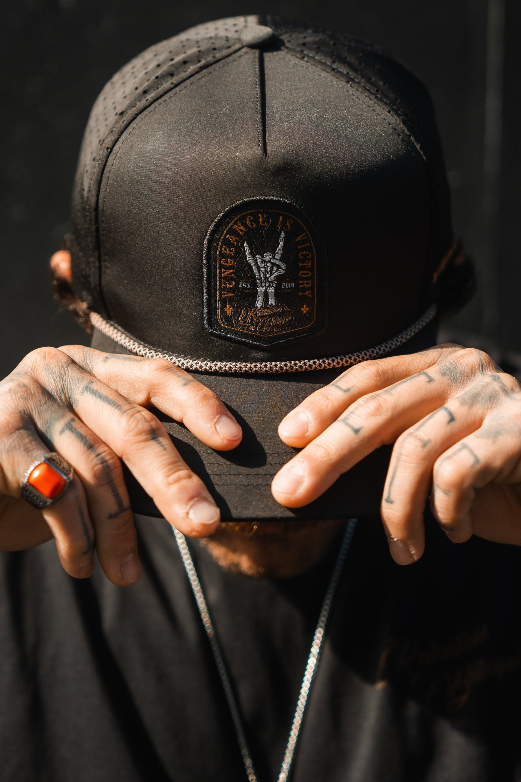 A person wearing the Vengeance Snapback by Relentless Betrayal lowers their head, partially concealing their face with tattooed hands—a subtle emblem of resilience. A red stone ring and a chain necklace complement the dark backdrop, whispering empowerment in silence.