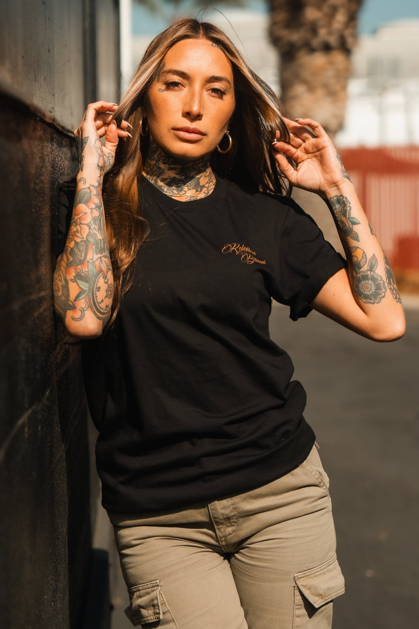 A person with long hair and tattoos leans against an outdoor wall, wearing a Vengeance Tee by Relentless Betrayal that signifies resilience. Sunlight highlights their face as a palm tree sways gently amidst blurred urban elements in the background.
