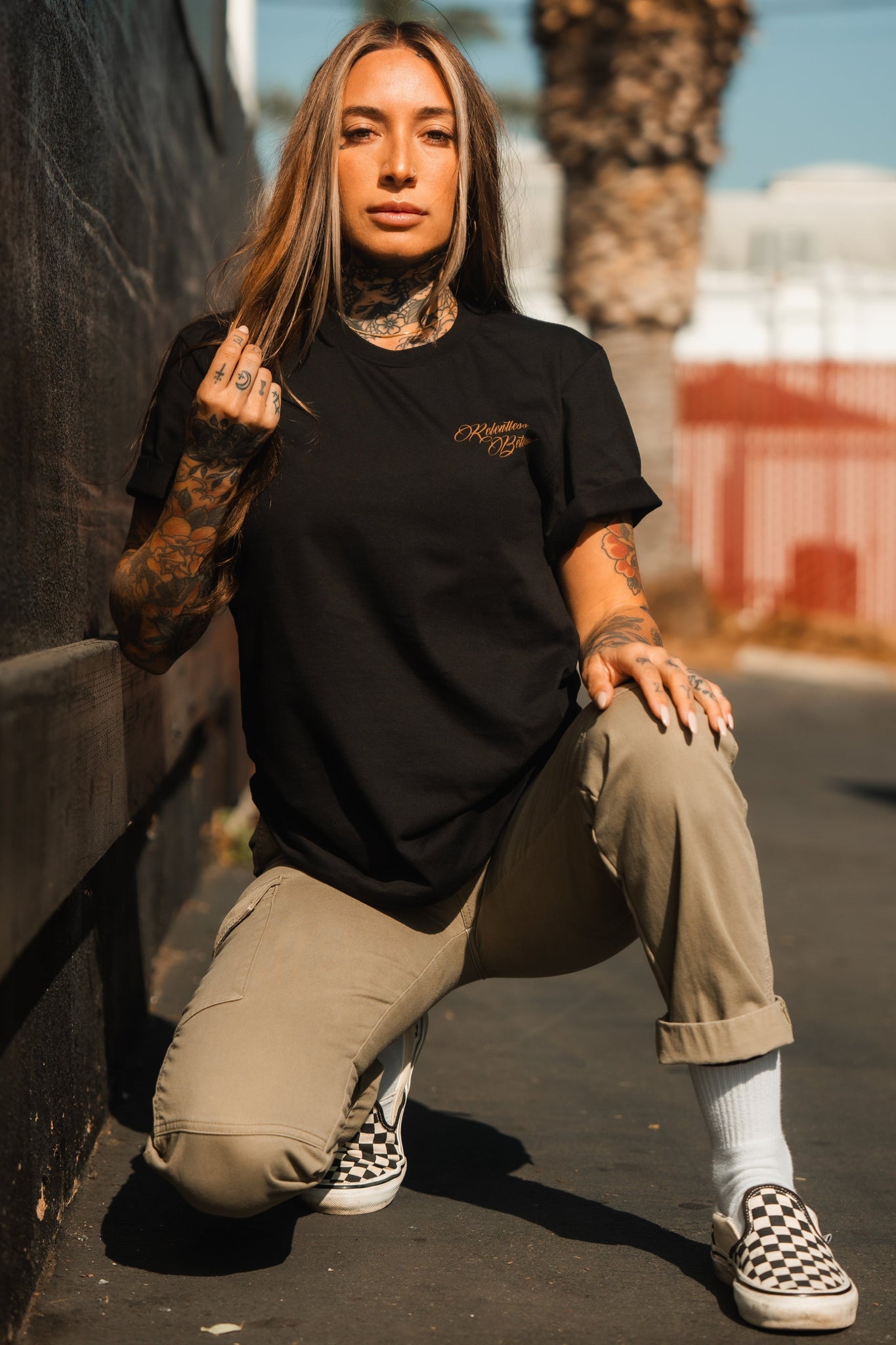 A person with long hair and tattoos kneels outside, radiating resilience and determination. They wear the Relentless Betrayal Vengeance Tee, beige pants, and checkered slip-ons against a blurred background of trees and buildings.