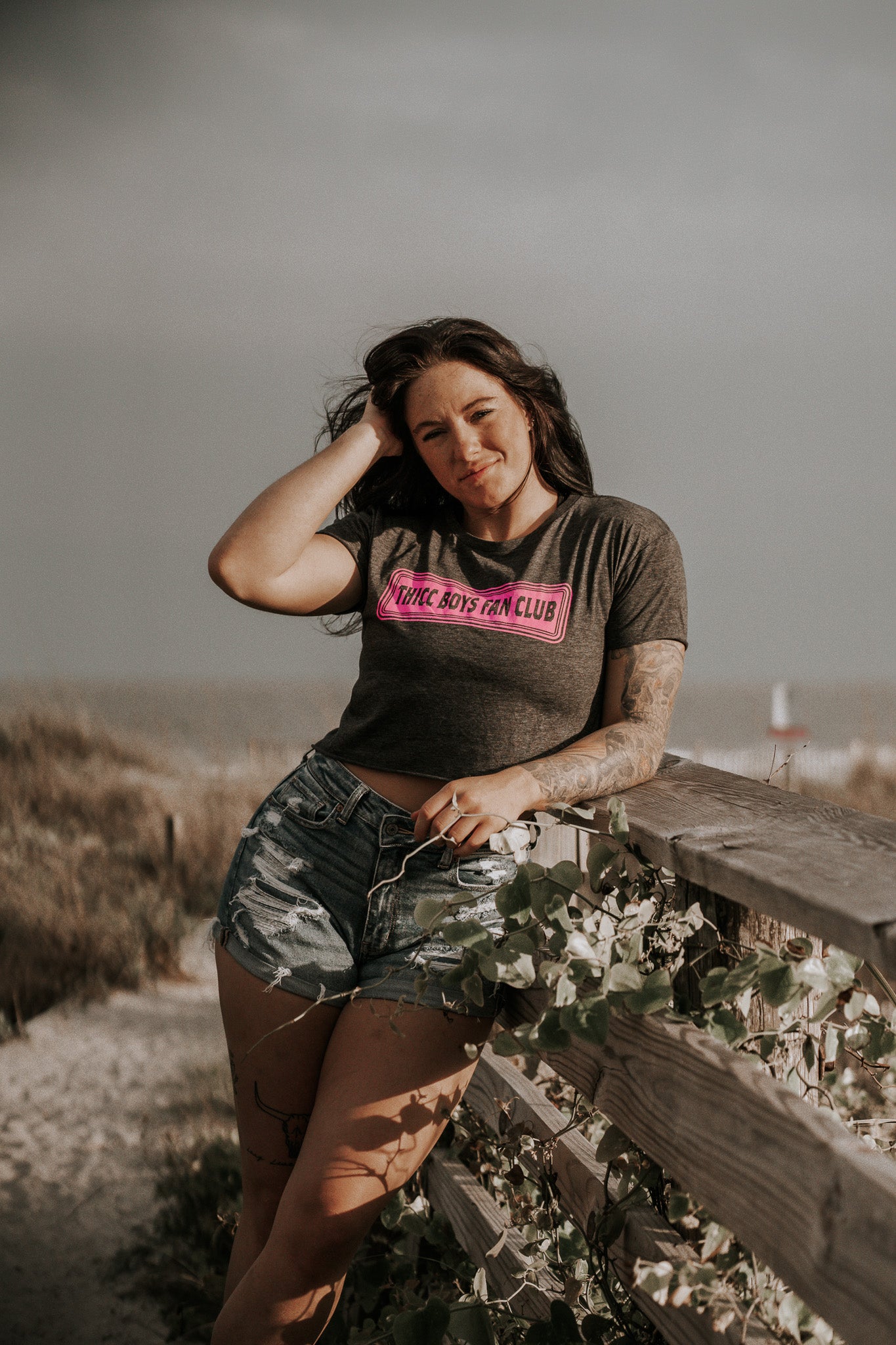 A woman with long dark hair, wearing a Tango Charlie Apparel THICC Boys Fan Club Crop T-Shirt in Charcoal and denim shorts, stands by a wooden fence at the beach with plants and sand in the background, looking at the camera and touching her hair.