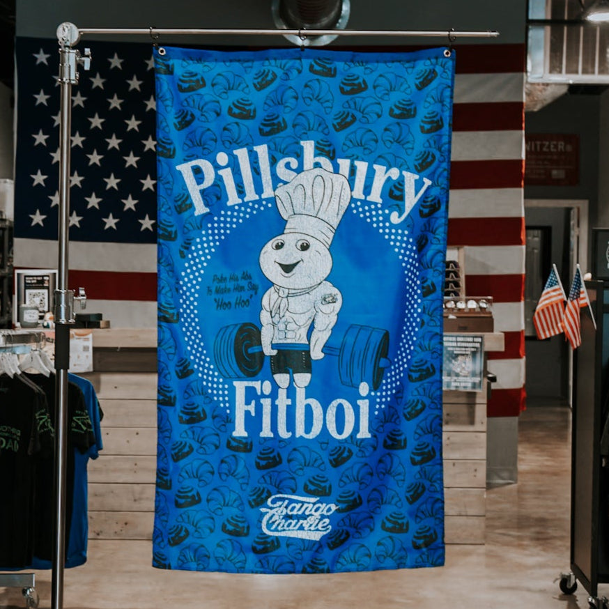 The Pillsbury Fitboi - Flag by Tango Charlie Apparel shows the Pillsbury Doughboy lifting a barbell on a blue flag, with Pillsbury Fitboi text. Ideal gym decor, this polyester flag adds unique style to your space.