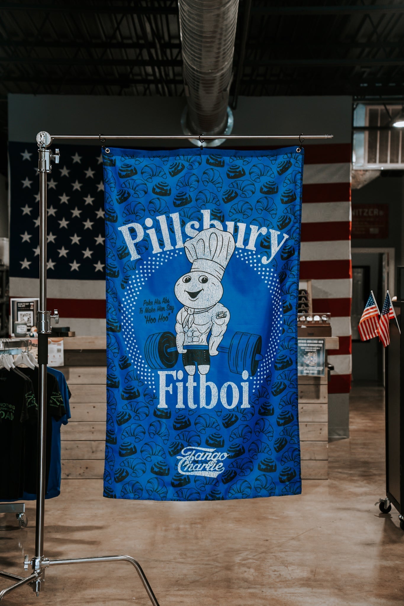 A blue banner labeled Pillsbury Fitboi by Tango Charlie Apparel shows a muscular cartoon doughboy in a chef hat lifting a dumbbell. A polyester flag, styled as an American flag, hangs in the industrial-style background.