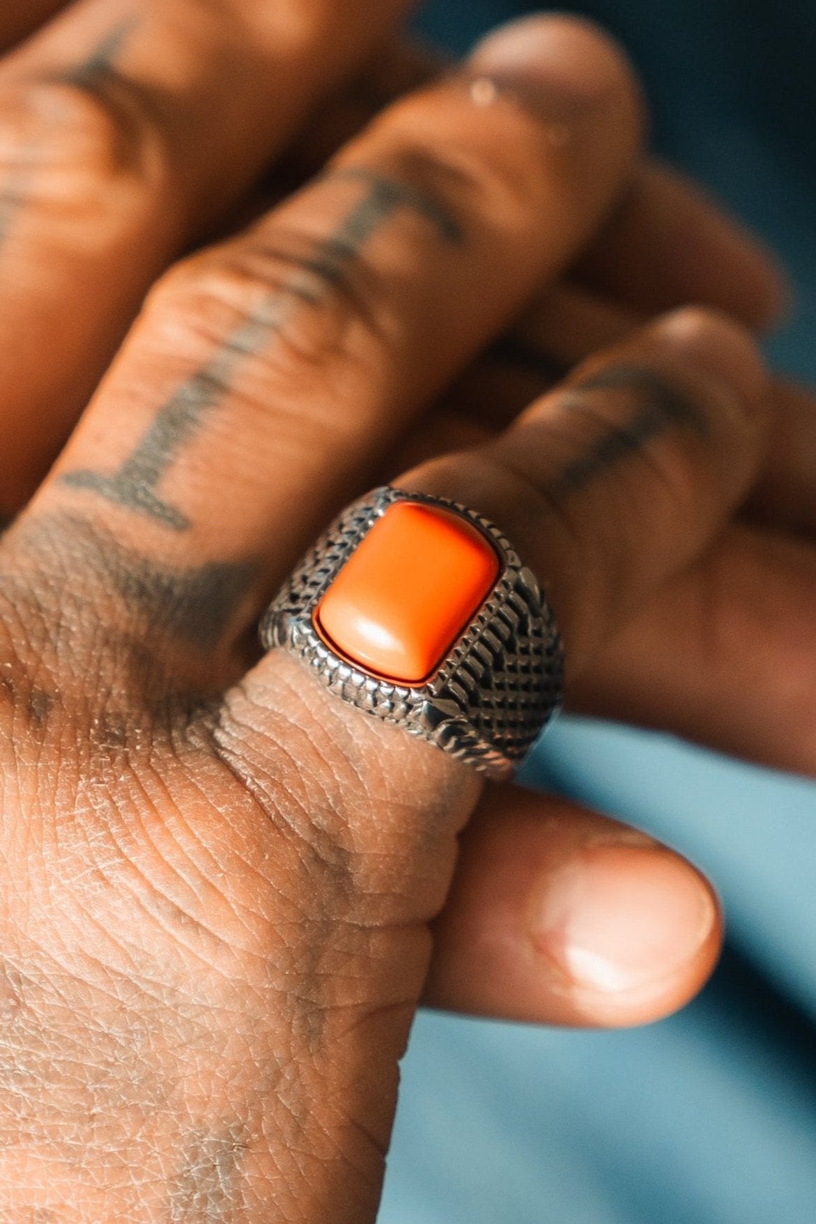 A tattooed hand showcases the Relentless Betrayal Hell Fire Ring, an ornate stainless steel piece with a large rectangular orange stone. The blurred background highlights the designs handcrafted elegance.