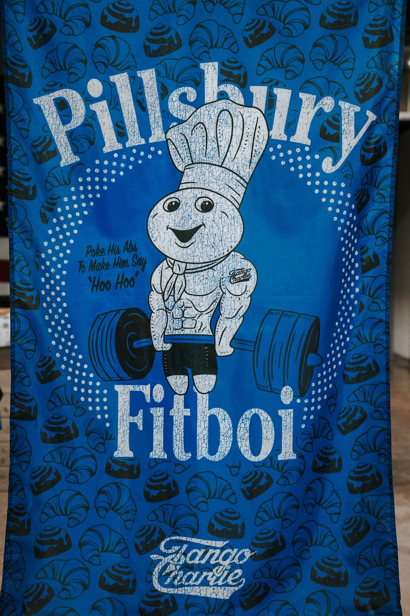 The Pillsbury Fitboi - Flag by Tango Charlie Apparel is a blue polyester towel featuring the Pillsbury Doughboy as a bodybuilder lifting a barbell. Text says Pillsbury Fitboi and Poke His Abs To Make Him Say Hoo Hoo, with crescent rolls in back.