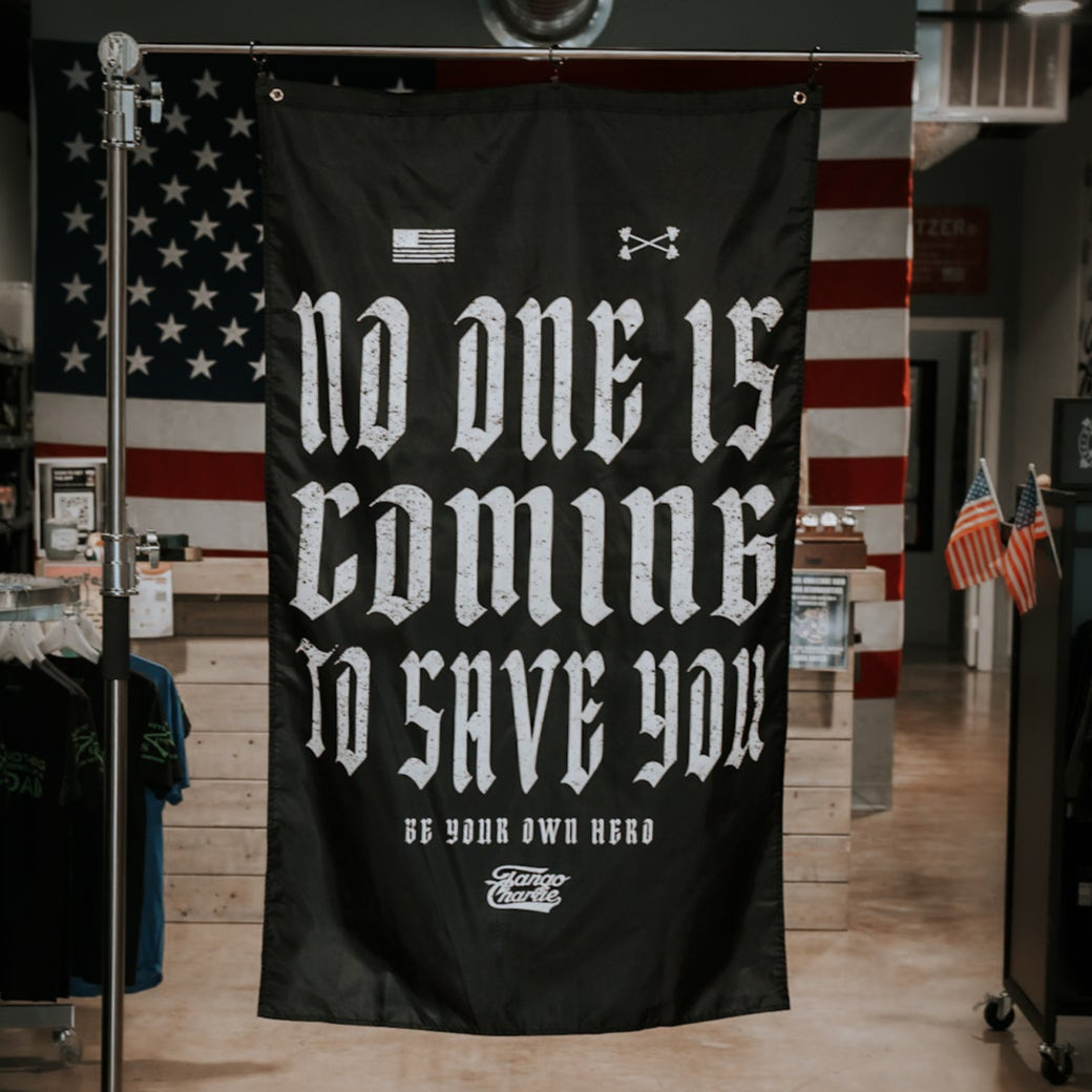 The No One Is Coming To Save You - Flag by Tango Charlie Apparel features bold white gothic text on black polyester, small American and Under Armour logos at the top, metal grommets for hanging, and an American flag in the background.