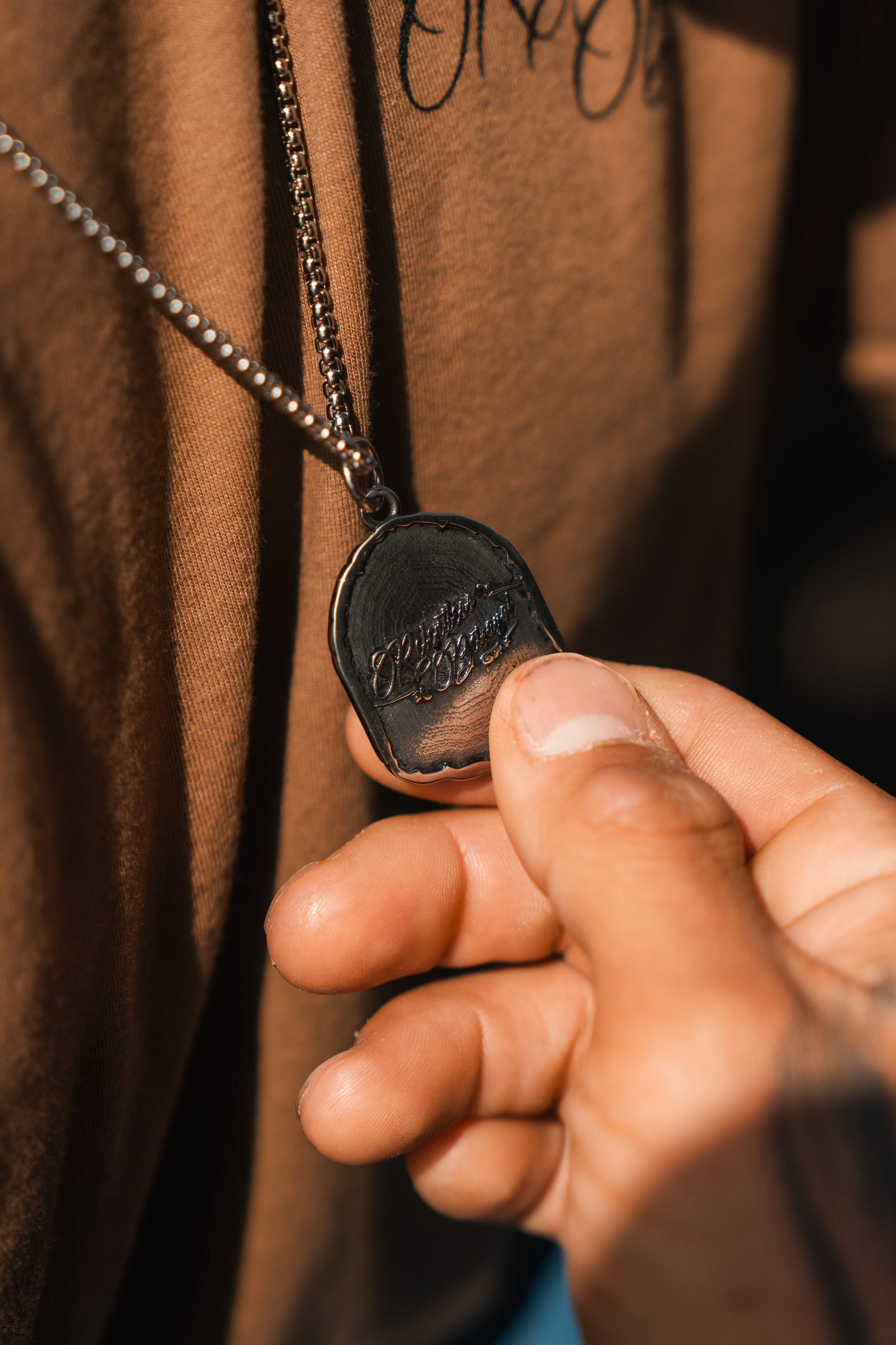 A close-up shows a hand holding the Vengeance Necklace by Relentless Betrayal, engraved with Vengeance is Victory, hanging from a chain. The wearer, in a brown shirt, embodies resilience and strength.