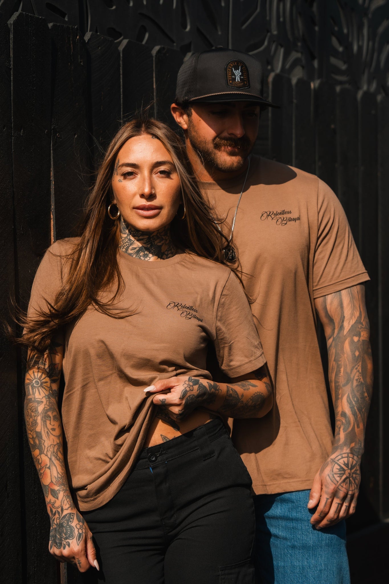 A tattooed woman and man stand side by side wearing matching Vengeance Camel T-Shirts from Relentless Betrayal and dark pants. The man wears a cap, and they are set against a dark background. Sunlight casts empowering shadows on their faces, embodying resilience in their stance.