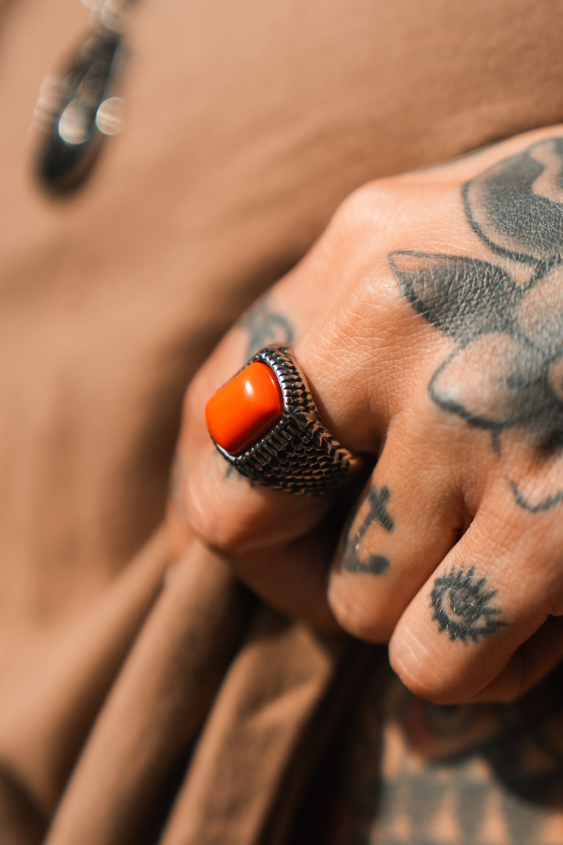 A tattooed hand, adorned with abstract and floral designs, showcases a handcrafted Hell Fire Ring by Relentless Betrayal, featuring a vibrant orange stone set in stainless steel against a soft brown fabric backdrop.