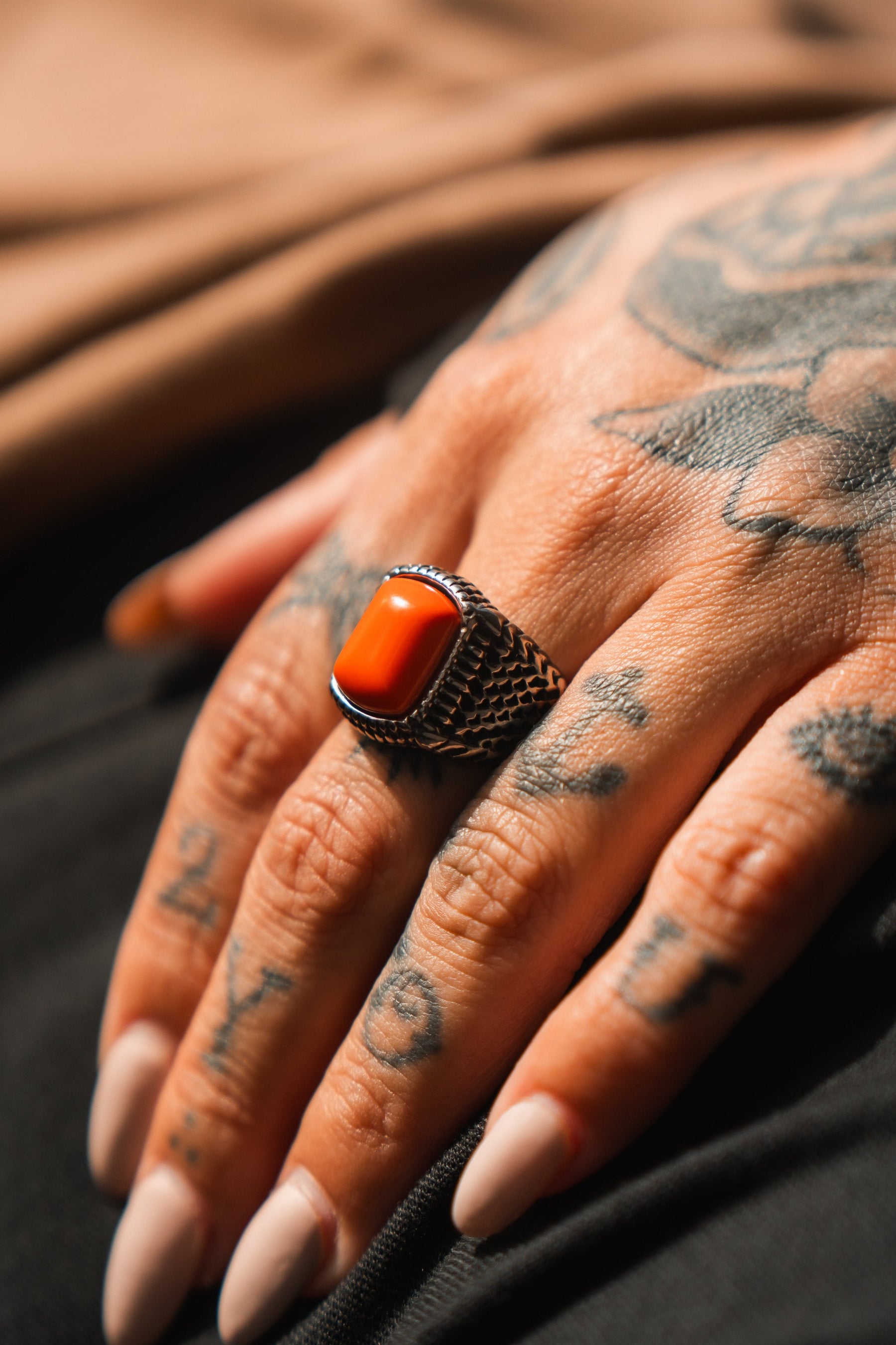 A tattooed hand with long nails displays the Relentless Betrayal Hell Fire Ring, featuring a vivid orange rectangular stone set in a detailed silver band.