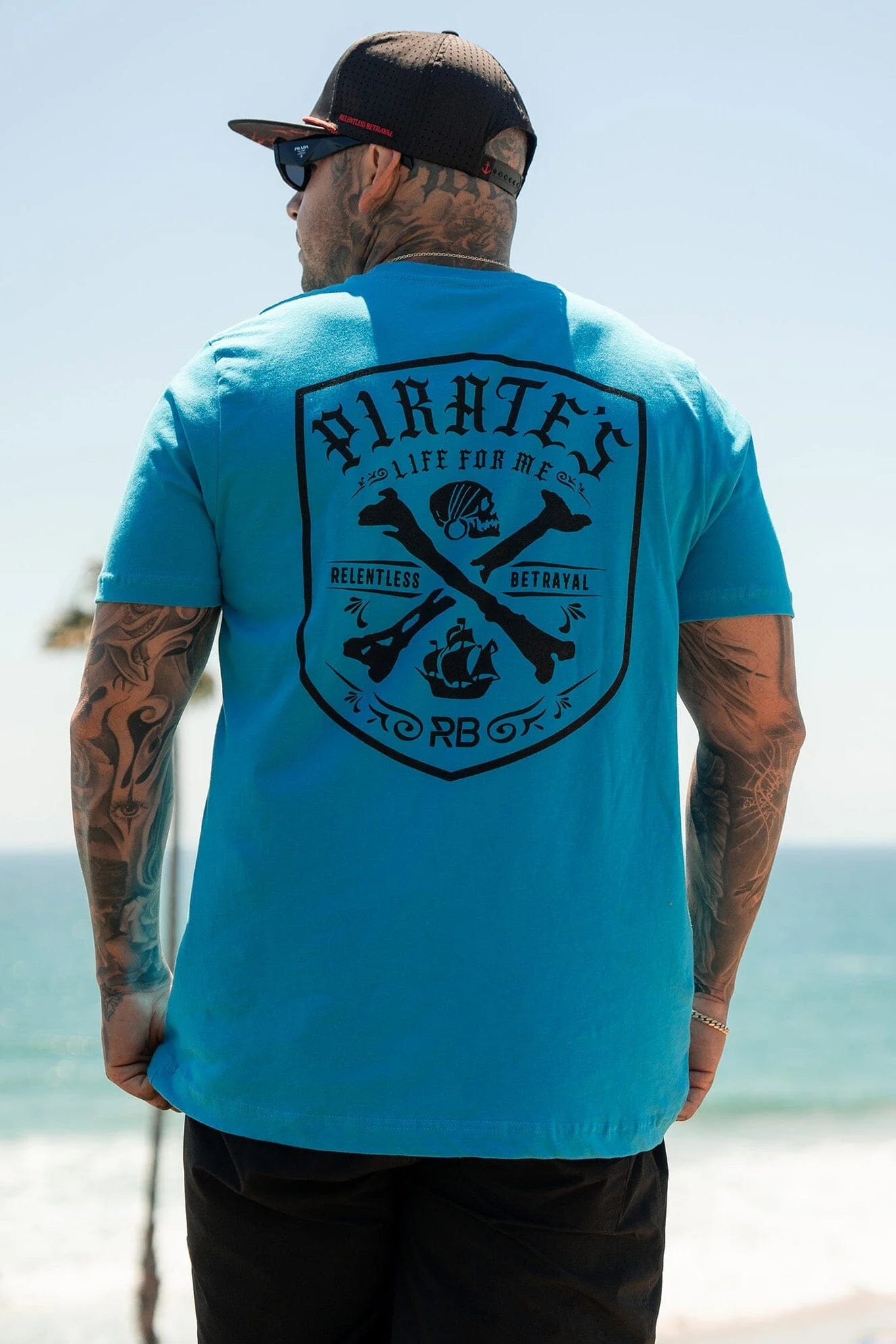 A tattooed man by the ocean dons a Pirates Life Aqua T-Shirt featuring black graphics of skulls, crossed swords, and Relentless Betrayal, embodying an outlaw’s freedom. He completes his look with sunglasses and a cap.