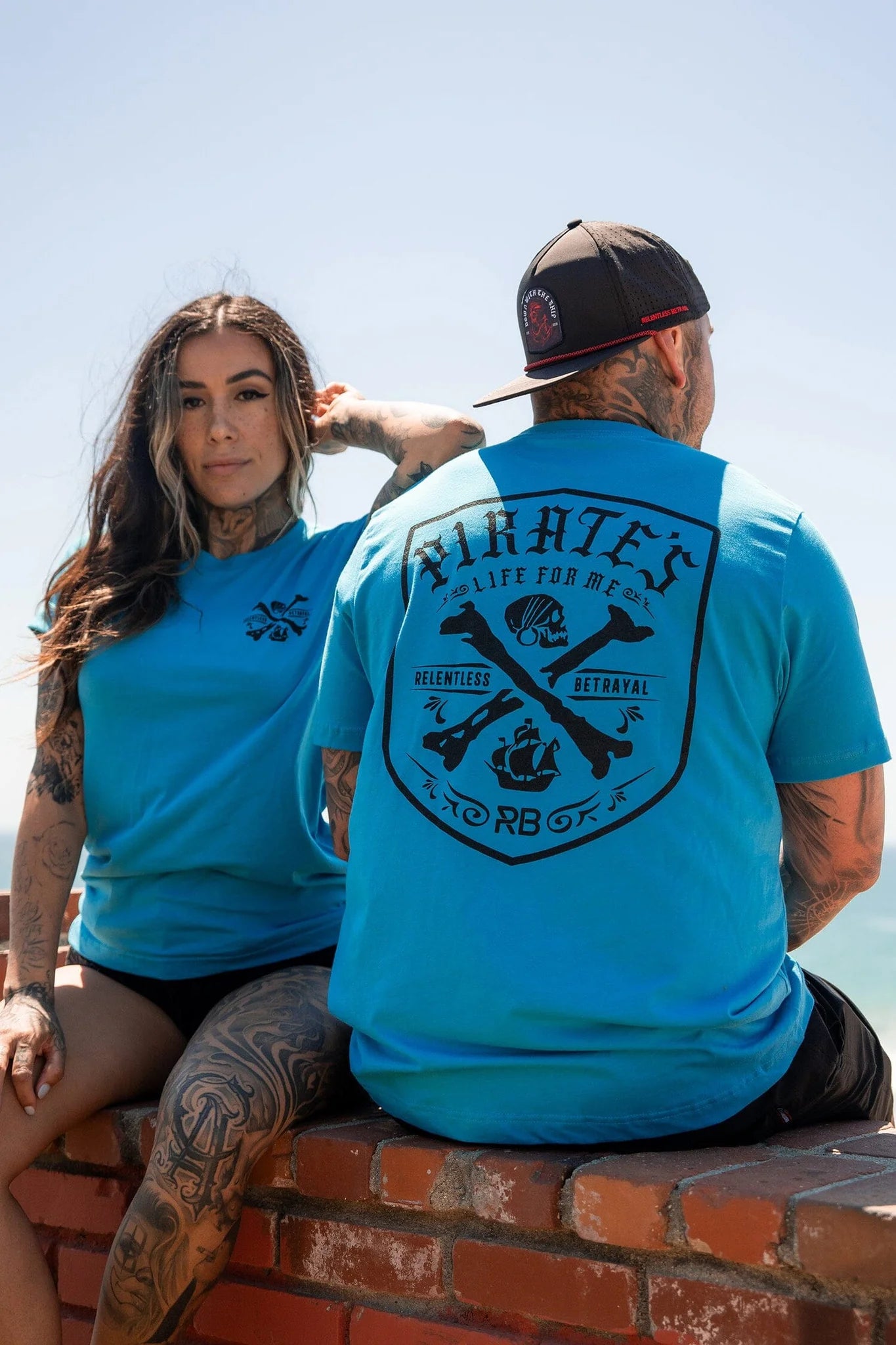 A couple sports matching Pirates Life Aqua T-Shirts from Relentless Betrayal. The woman perches on a brick wall; the man, donning a cap with his back to the camera, stands nearby. Both reveal tattoos against a vivid sky backdrop.