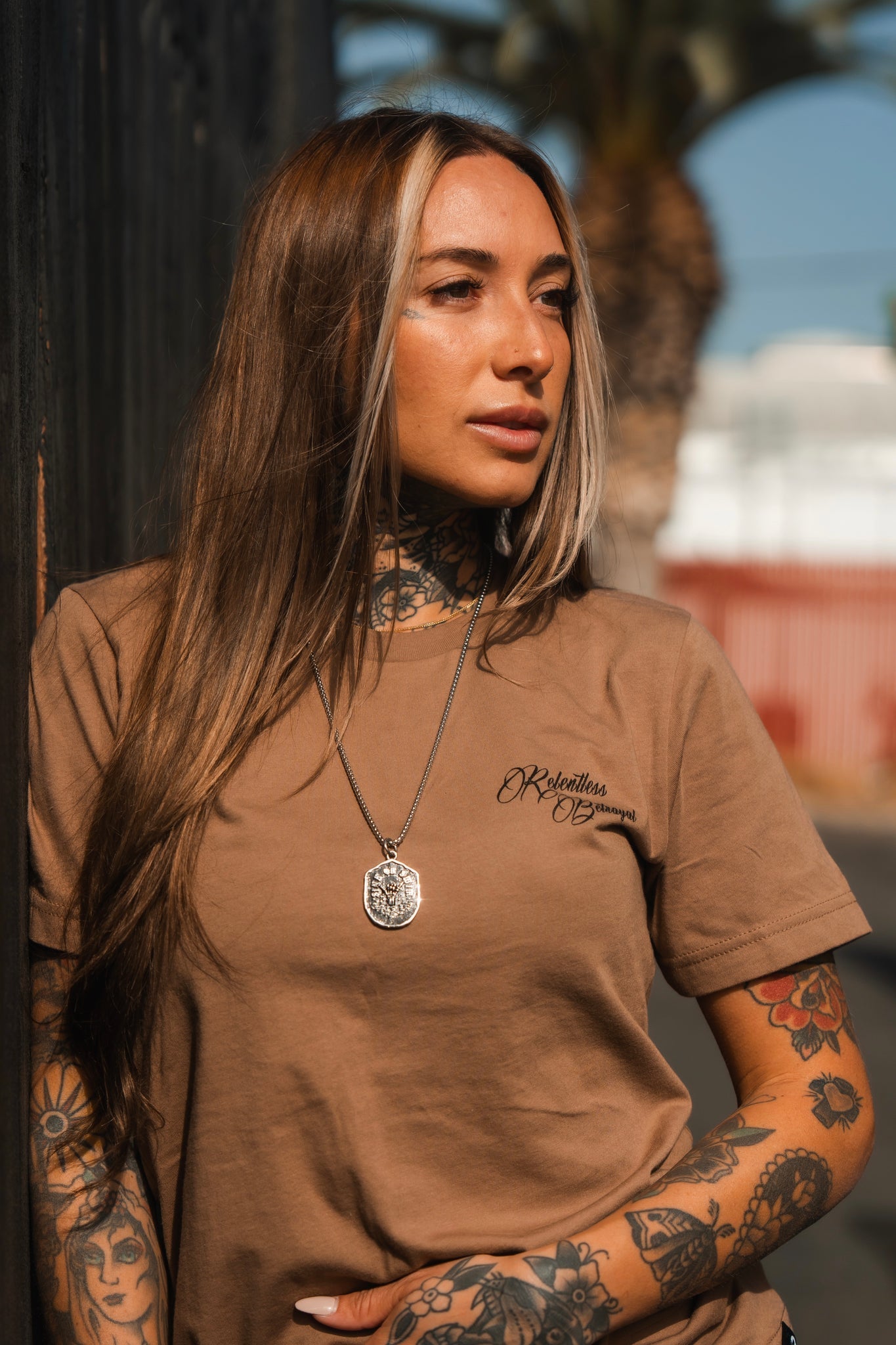 A tattooed woman with long hair leans against a fence, her gaze reflecting resilience. She wears a Vengeance Camel T-Shirt by Relentless Betrayal and a pendant necklace. Palm trees and a hazy building enhance the empowering backdrop of her stance.