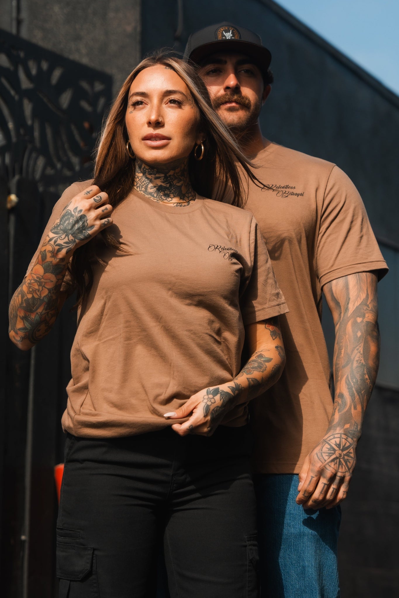 Two tattooed individuals stand confidently outdoors, embodying resilience. The woman wears black pants and a Vengeance Camel T-Shirt by Relentless Betrayal. Behind her, a man matches in brown with a black cap. Both proudly display intricate tattoos on their arms and necks.