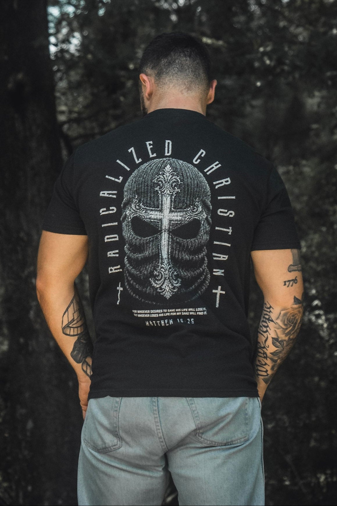 A man stands outdoors with his back to the camera, wearing the 13th Radicalized Christian Premium Tee. The black t-shirt features a masked face with a cross and the words “Matthew 16:25,” boldly referencing the Bible verse: “For whoever wants to save his life will lose it.”.