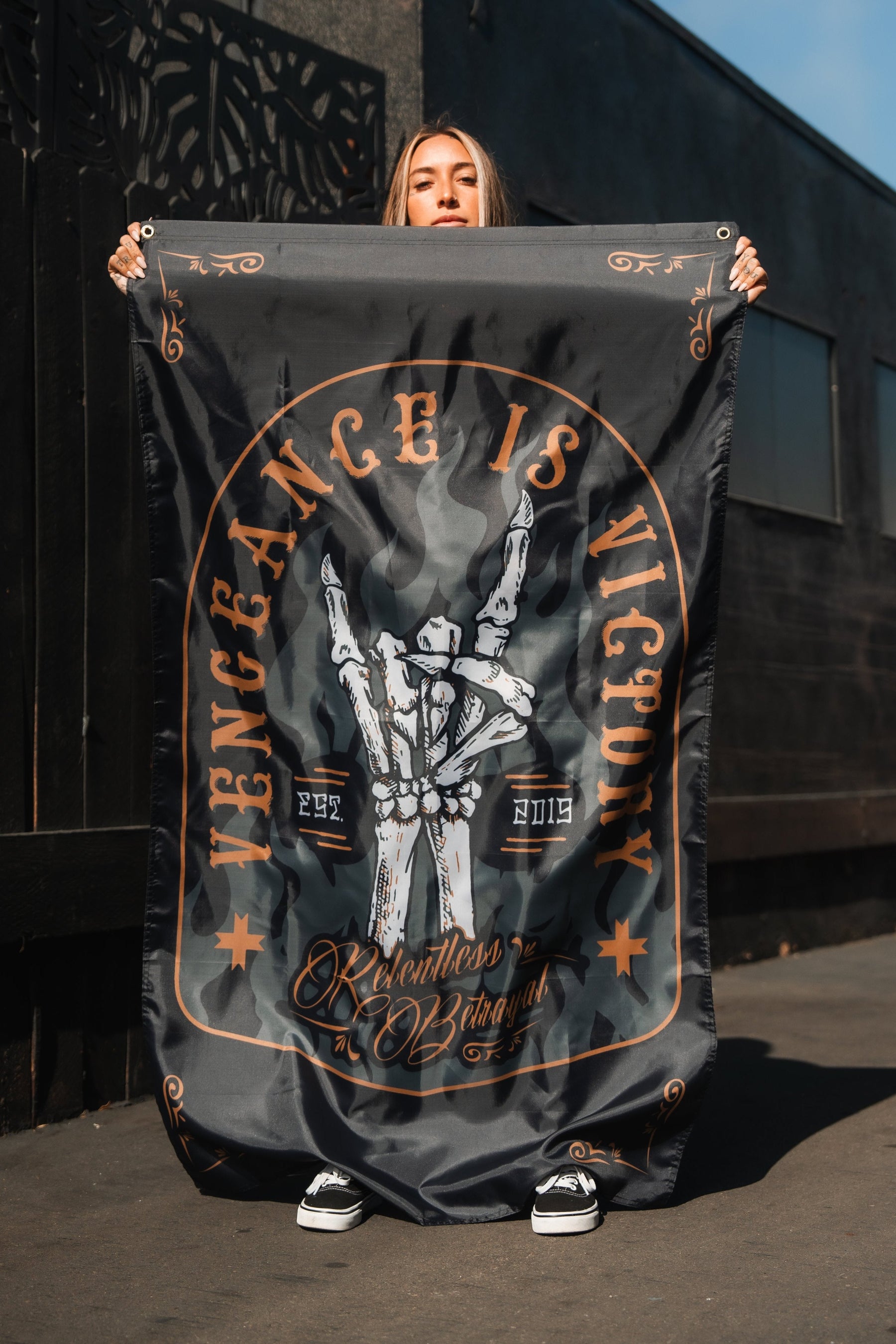 A person holds the Vengeance Flag from Relentless Betrayal, featuring skeletal hands in a peace sign with Vengeance is Victory, and the years 2022 and 2019, symbolizing empowerment, against a dark building under a clear sky.