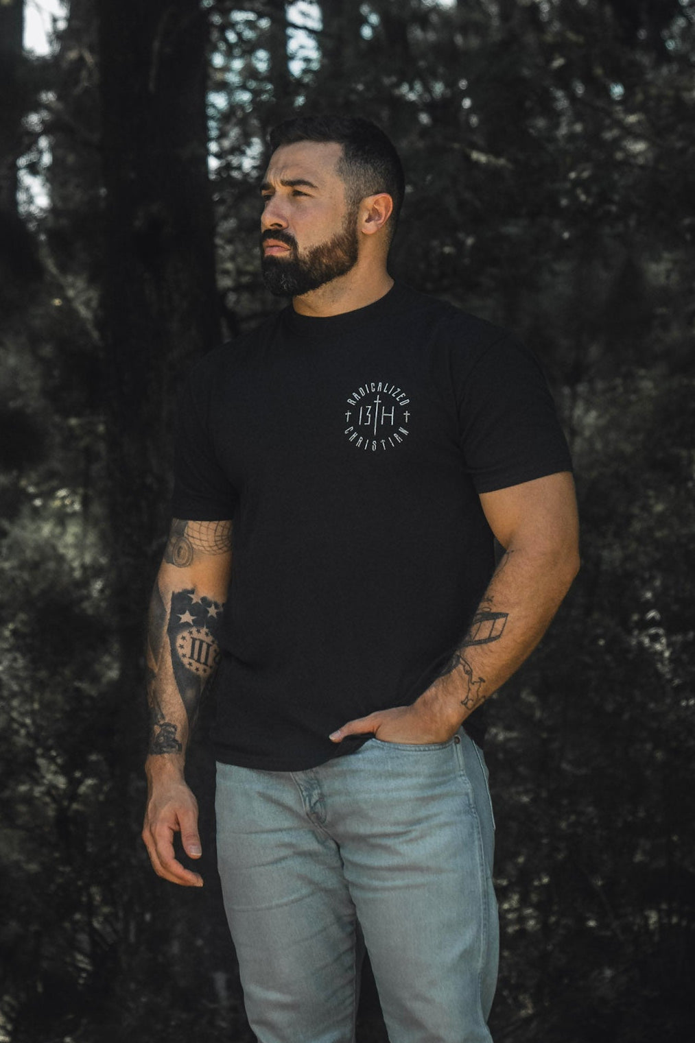 A tattooed, bearded man stands outdoors against a forest backdrop in dim natural light, dressed in the Radicalized Christian Premium Tee by 13th and light jeans. With one hand in his pocket and looking contemplatively to the side, he seems to reflect on the message of Matthew 16:25—a verse about discovering what it truly means to save your life.