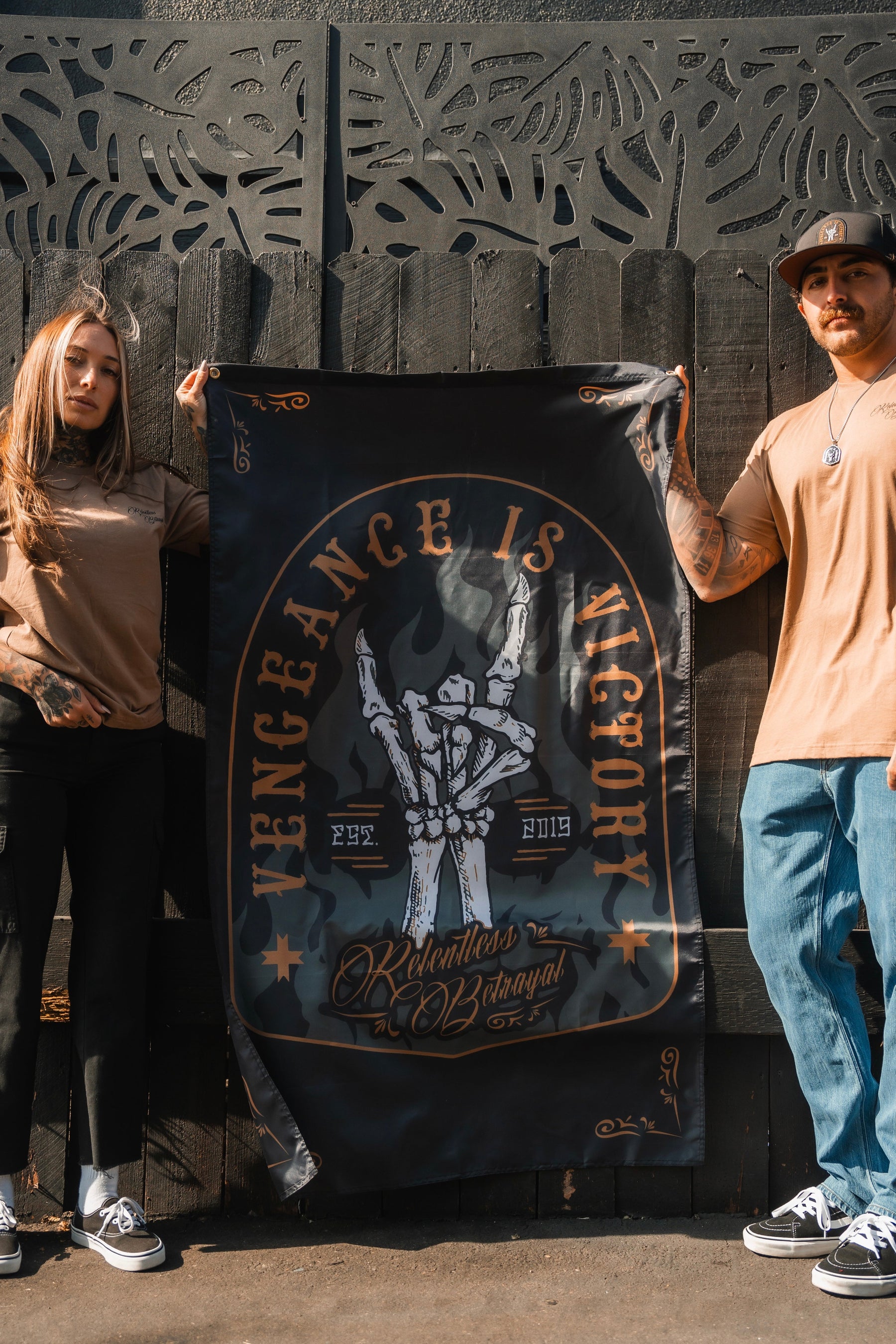 Two people hold the Relentless Betrayals Vengeance Flag, featuring a skull and Vengeance is Victory against a wooden fence. Dressed casually, they exude resilience as soft shadows play in the sunlight.