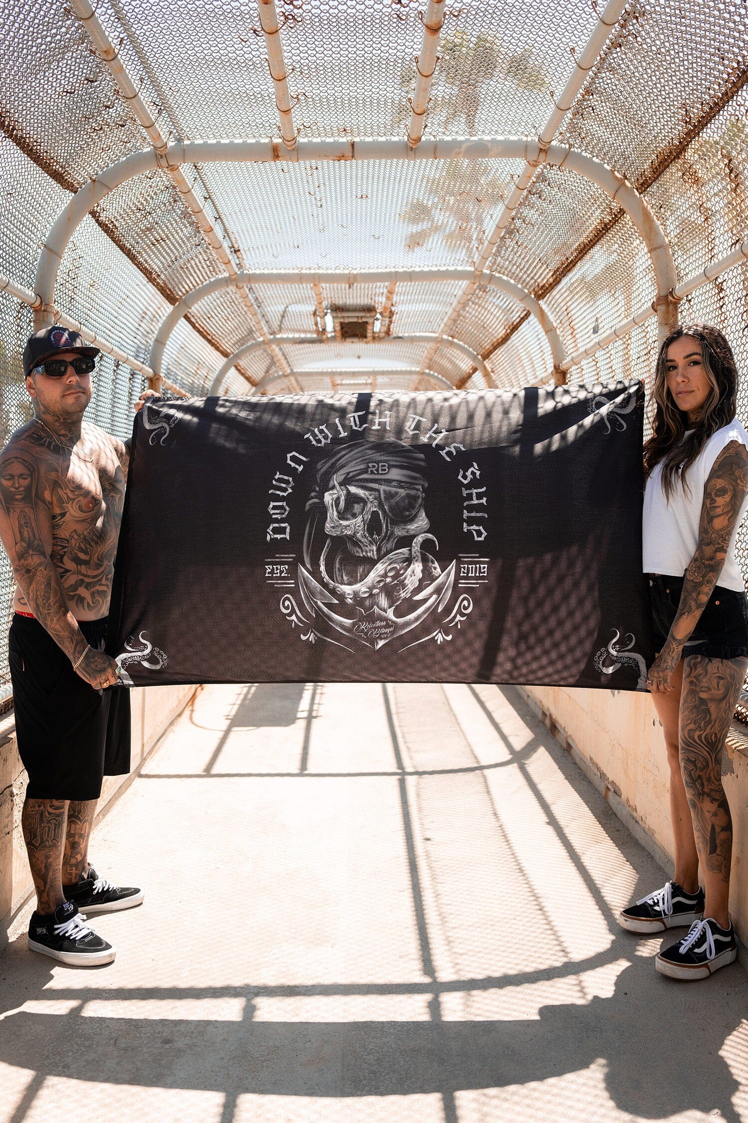 Two tattooed individuals stand on a bridge with metal mesh, holding the Down With the Ship Flag from Relentless Betrayal. Made of durable polyester, the pirate-themed flag features skull and nautical elements. Both wear casual clothing and sunglasses as shadows are cast on the ground.