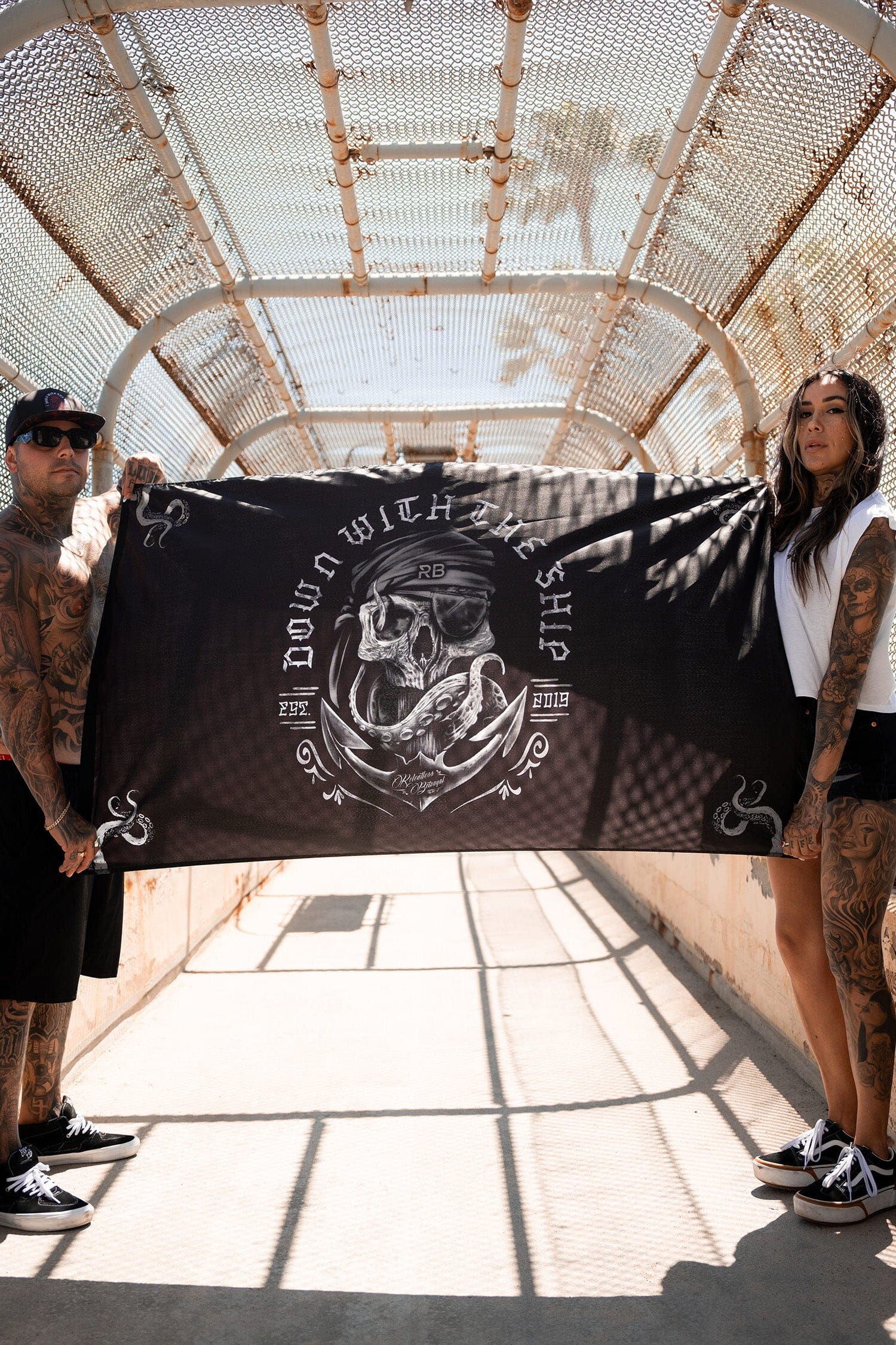 Two tattooed individuals hold the Down With the Ship flag by Relentless Betrayal, crafted from durable polyester with a skull and nautical design. They stand under sunlight on a pedestrian bridge, and the flags metal grommets glint softly in the light.