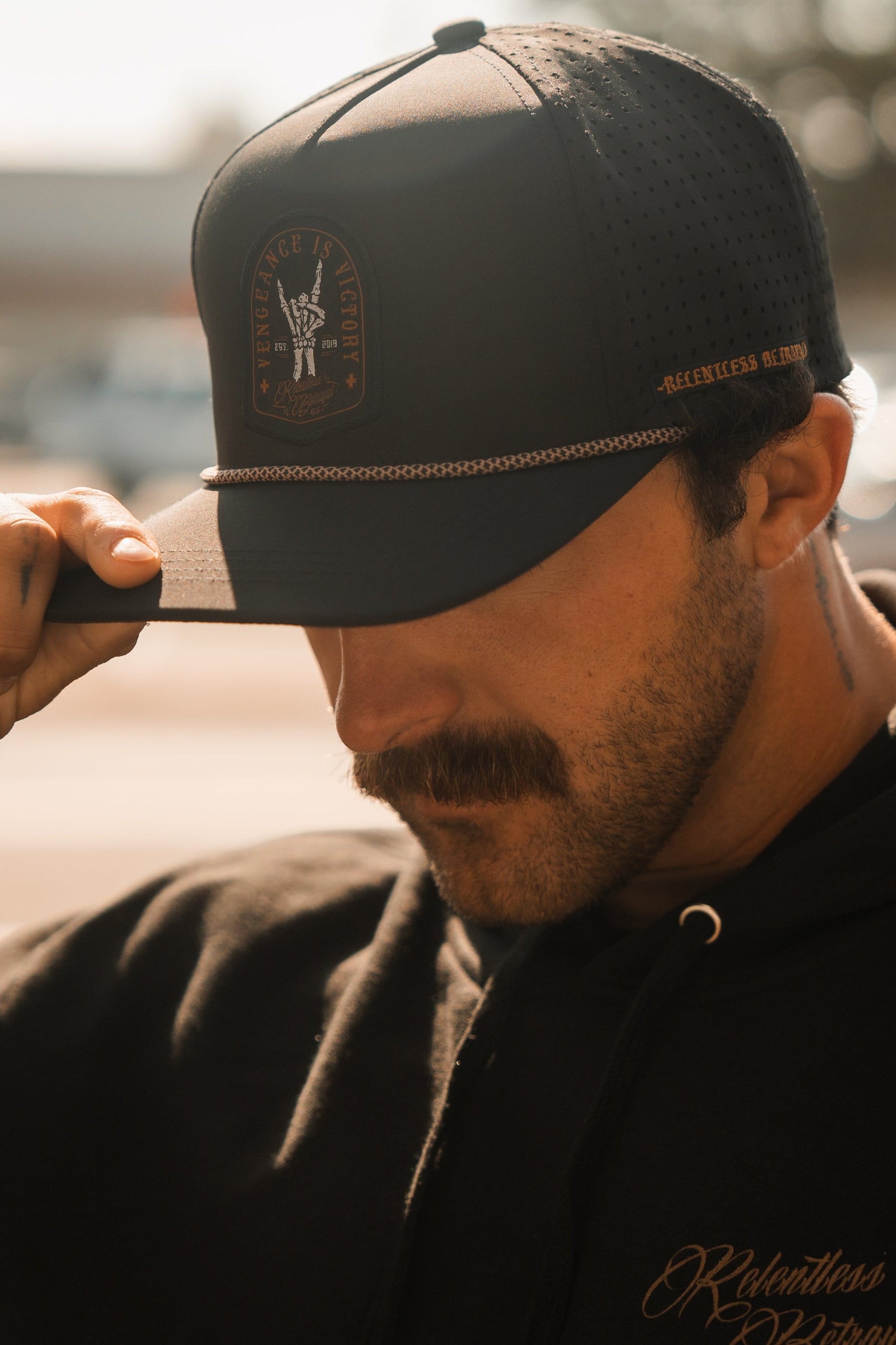 A resilient man with a mustache wears a Relentless Betrayals Vengeance Snapback and hoodie, tilting the brim of his cap downward against a softly blurred, sunlit background.