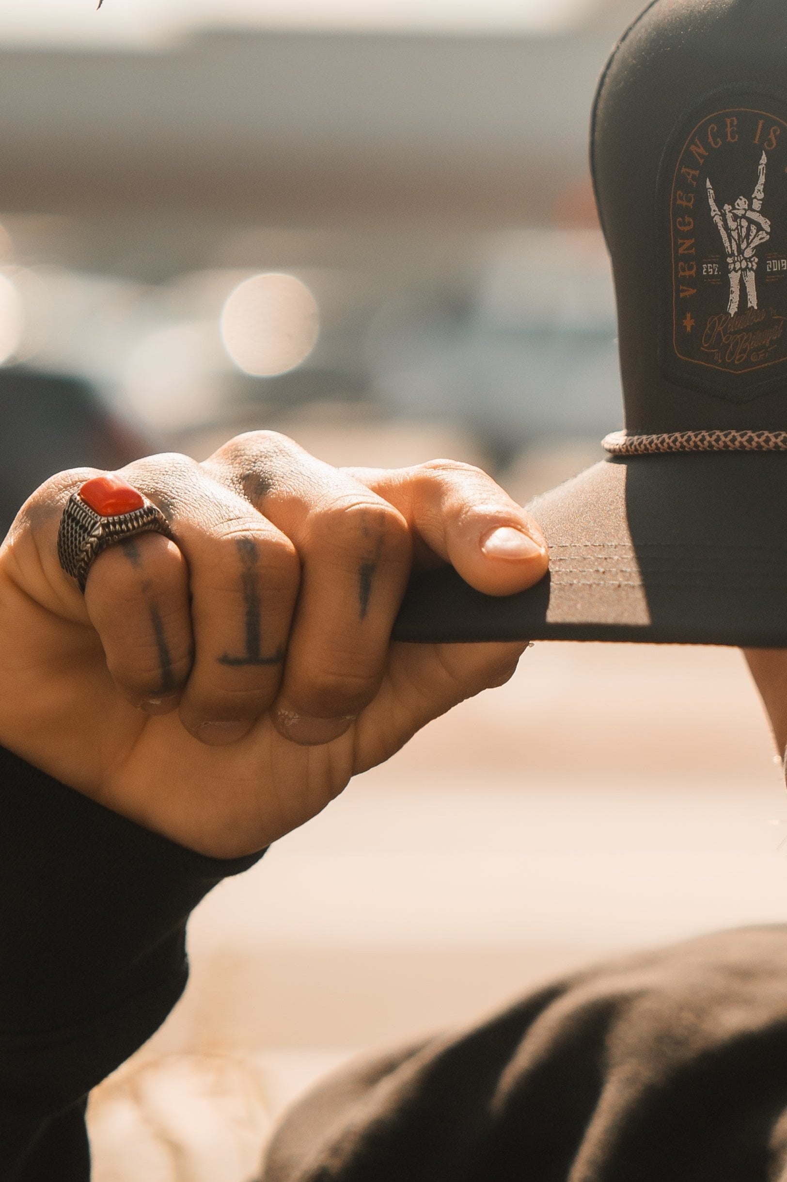 Tattooed fingers with a Relentless Betrayal Hell Fire Ring grip a baseball cap featuring a sword patch and Vengeance Is text. Sunlight highlights the ring and hand against a blurred background.