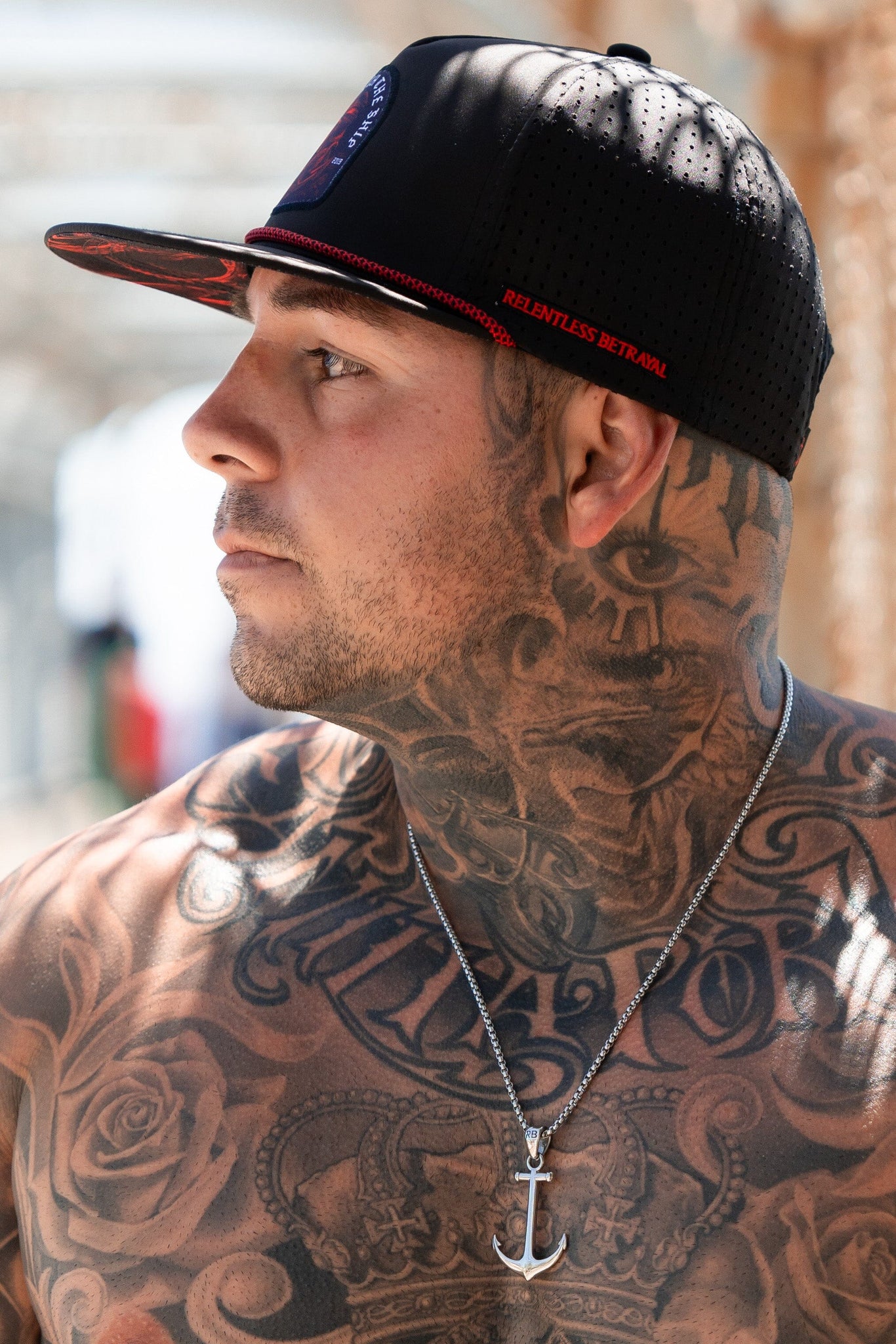 A man with floral and text tattoos on his neck and chest wears a red Ship Wreck Snapback by Relentless Betrayal and a silver anchor necklace, gazing sideways in an outdoor setting.