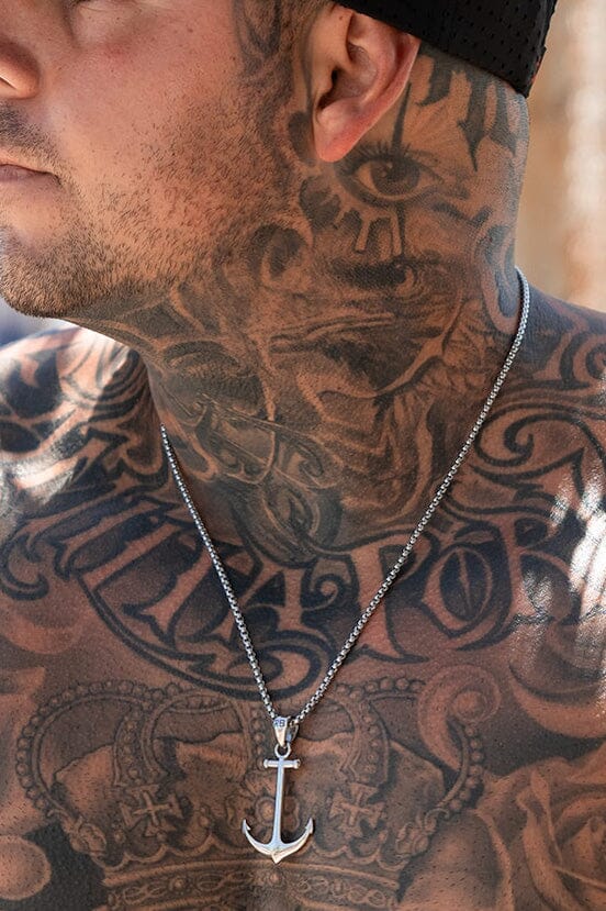 A man with extensive tattoos on his neck and chest wears a black cap, adorned with the Anchor Drop Necklace by Relentless Betrayal—an ornate 26-inch handcrafted stainless steel chain featuring an anchor pendant.