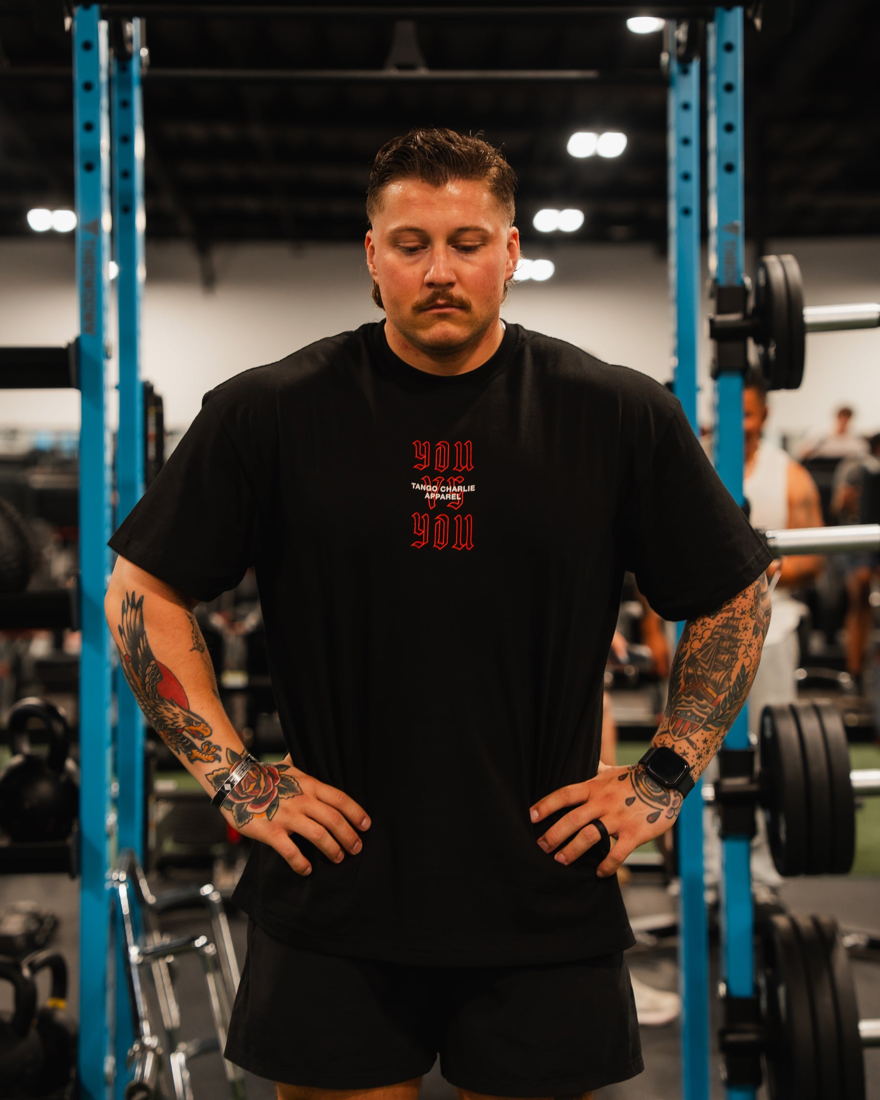 A tattooed man stands in a gym, hands on hips, wearing the You vs You - Premium Oversized Tee from Tango Charlie Apparel. Weightlifting equipment and other people are visible in the background.