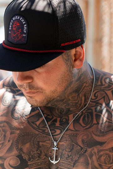 A tattooed man wears a black cap with red trim, featuring a patch and Relentless Better text from Relentless Betrayal. An Anchor Drop Necklace with a 26-inch handcrafted chain hangs around his neck, and the caps shadow partly conceals his face.