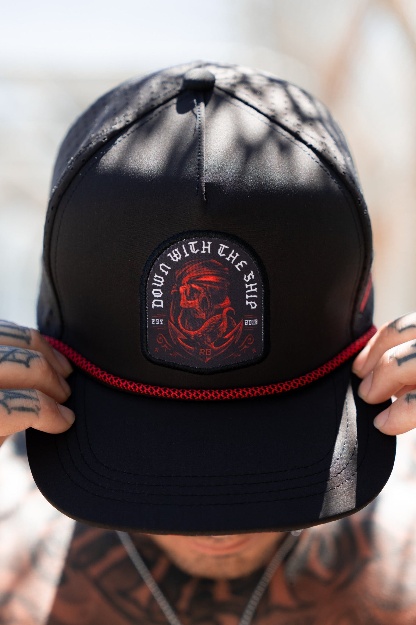 A person wearing the Ship Wreck Snapback - Red by Relentless Betrayal, featuring a skull design and DOWN WITH THE SHIP patch with a red braided strap, adjusts the cap, displaying tattoos on their fingers.