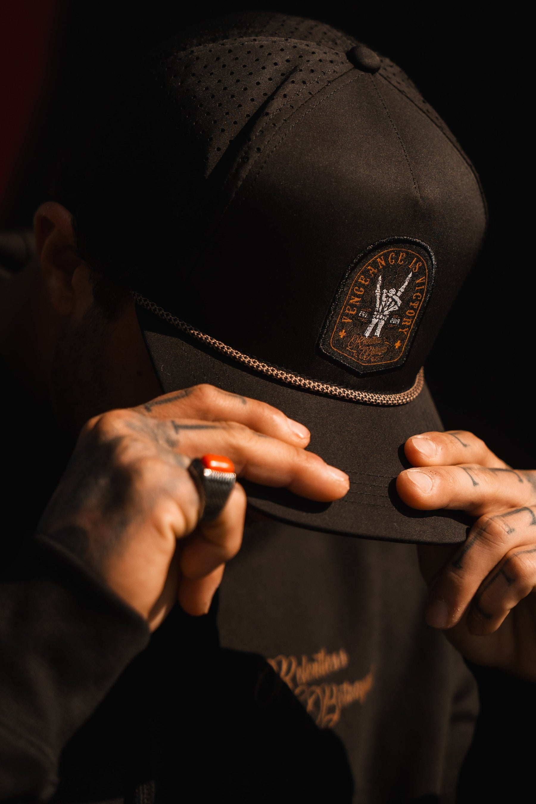 A person in a Relentless Betrayal Vengeance Snapback adjusts the brim with tattooed fingers, showing resilience. Beneath a black jacket, shadows obscure their face.
