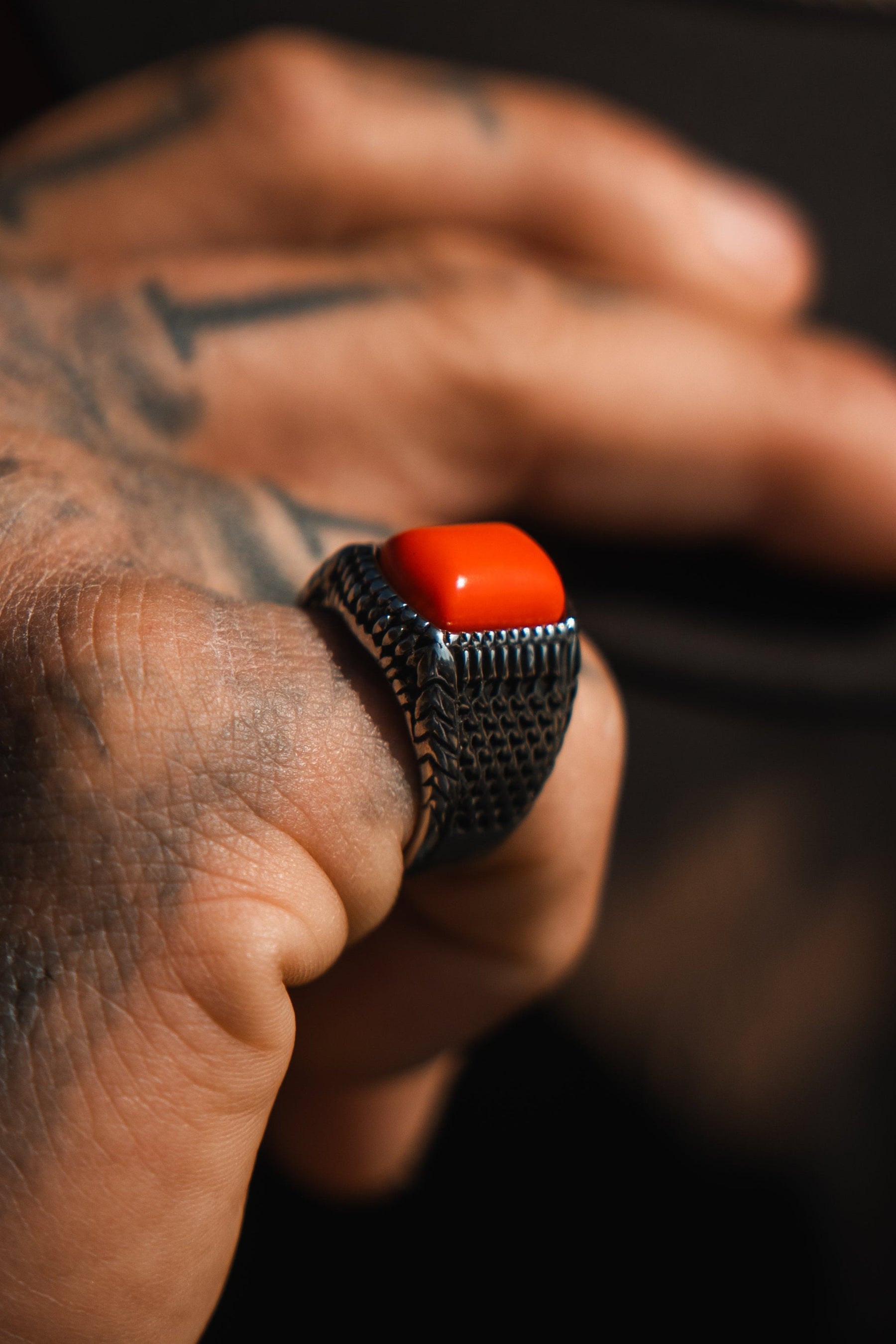 A tattooed hand dons Relentless Betrayals handcrafted Hell Fire Ring, showcasing a large red stone centerpiece. Its textured silver encircles the finger, resting on a dark surface and emphasizing the contrast between the vibrant stone and skin.