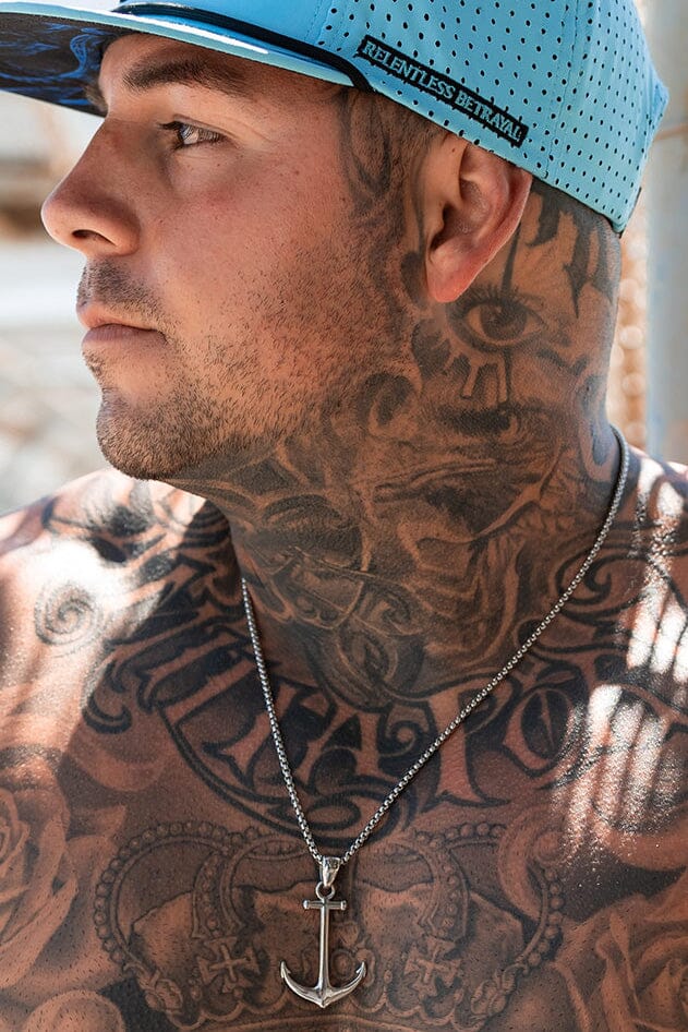 A tattooed man wearing a blue cap with Relentless Betrayal printed on it and a handcrafted Anchor Drop Necklace on a 26-inch stainless steel chain glances sideways.