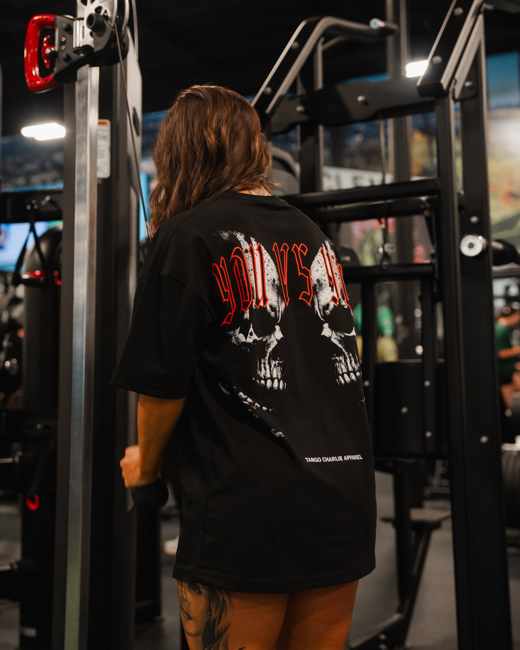 A person with long brown hair and tattooed legs stands in a gym, back to the camera, wearing the Tango Charlie Apparel You vs You Premium Oversized Tee featuring skull graphics and red lettering.