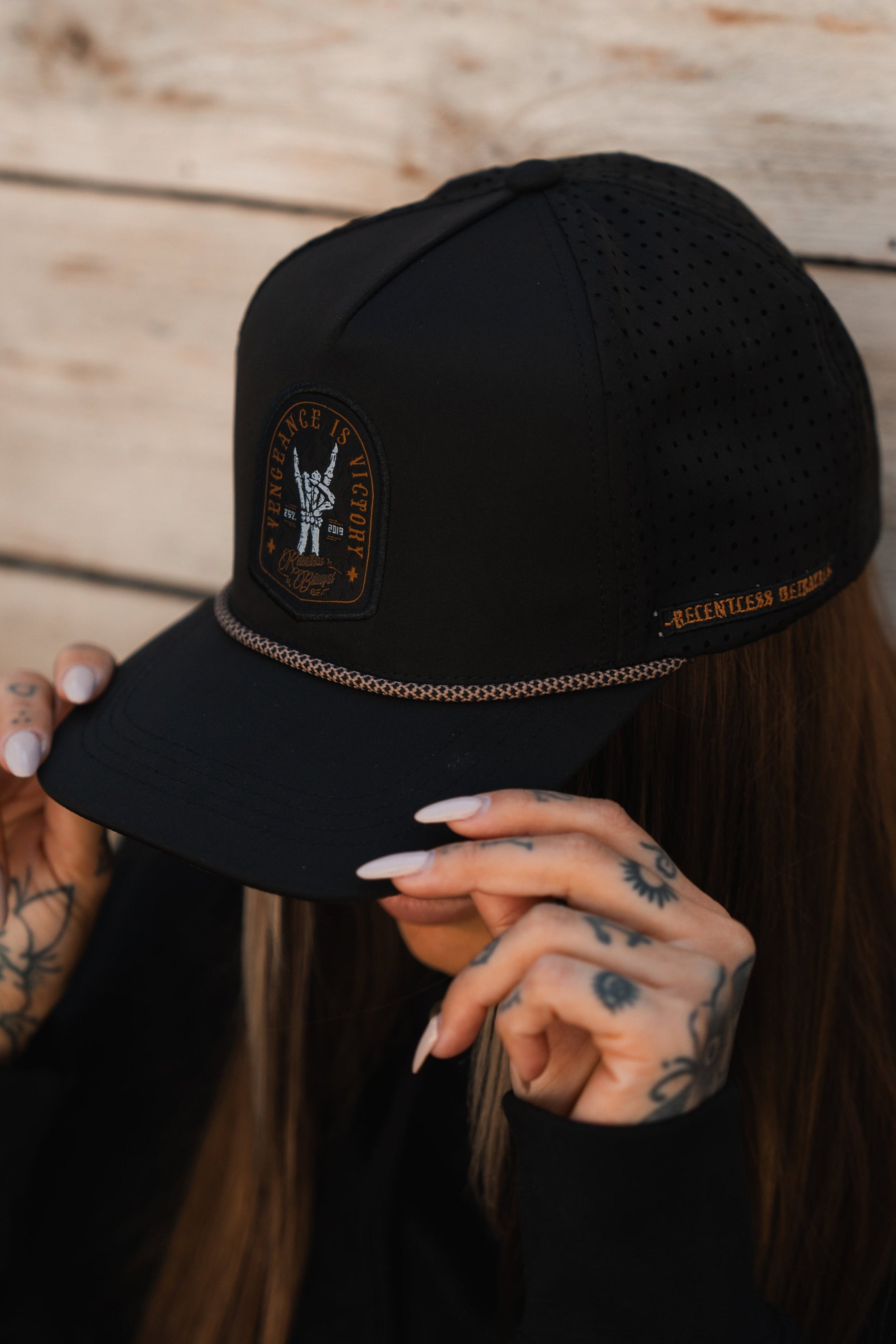 A person with tattooed hands adjusts their black Vengeance Snapback by Relentless Betrayal, featuring an eagle patch with Endurance 18 Eight and Relentless Retreat, and perforated sides, against a rustic wooden backdrop.
