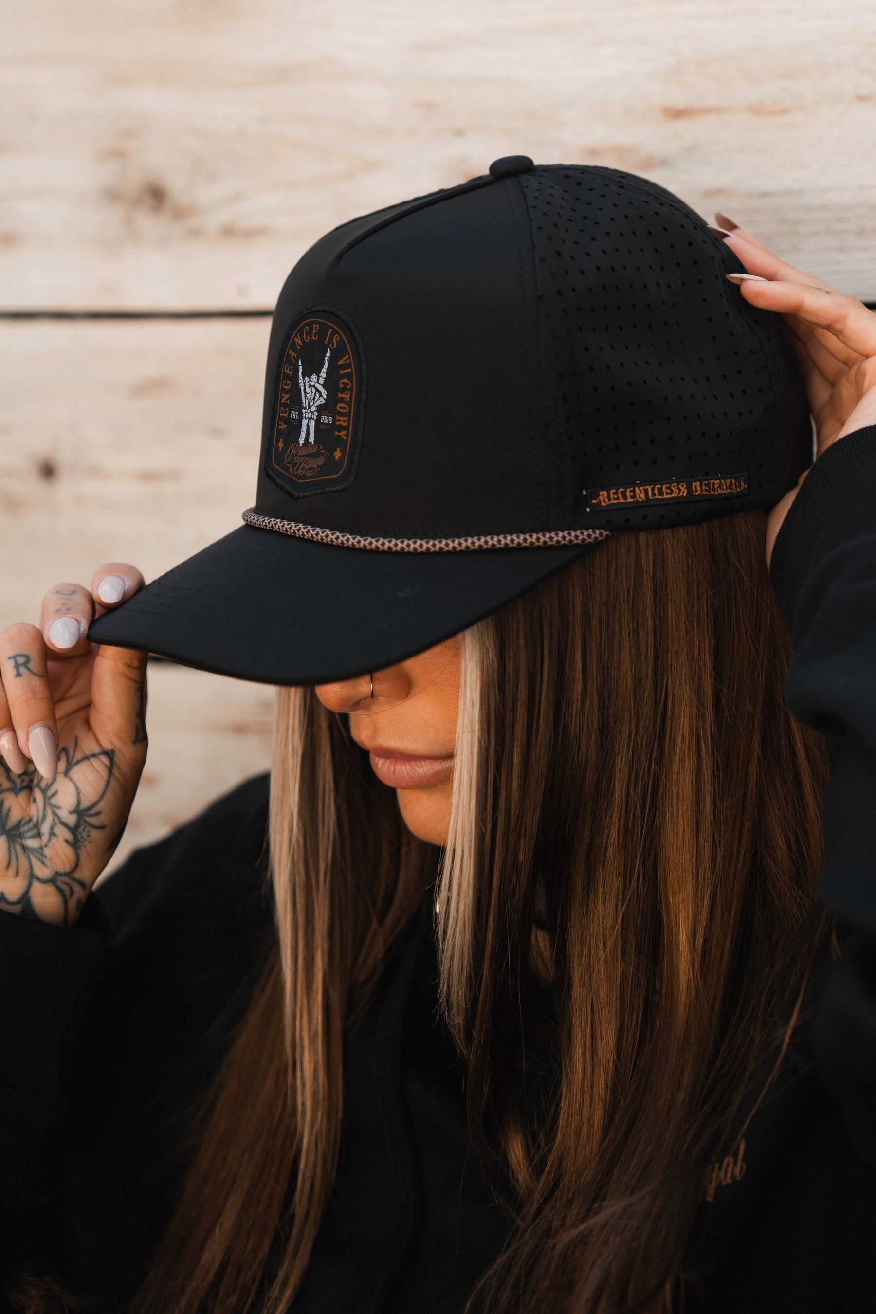 A person with long hair wearing the Relentless Betrayal Vengeance Snapback, a black cap with a Vengeance is Victory patch and chain, partially obscuring their face. Theyre clad in a black sweatshirt and display finger tattoos, posed against a wooden backdrop.