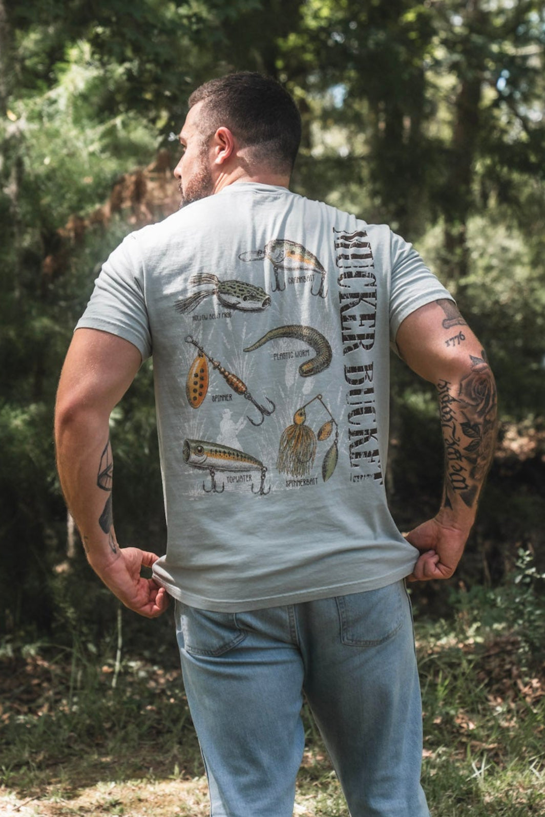 A man stands outdoors in a wooded area, wearing the Mucker Bucket Bait DNA Premium Tee—a light gray, premium relaxed fit shirt made from soft ring-spun cotton. Featuring fish illustrations and the words CATCH MORE on the back, he pairs it with light blue jeans.