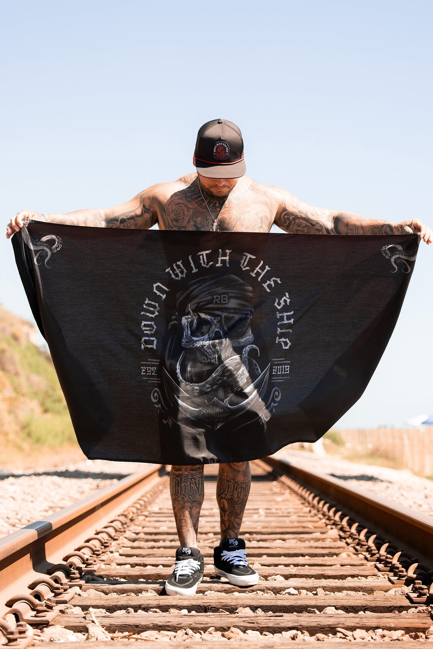 Standing on train tracks, a tattooed person in a cap showcases the Relentless Betrayal Down With The Ship Flag—a large black polyester piece with a skeleton design and metal grommets—set against clear skies and grassy hills.