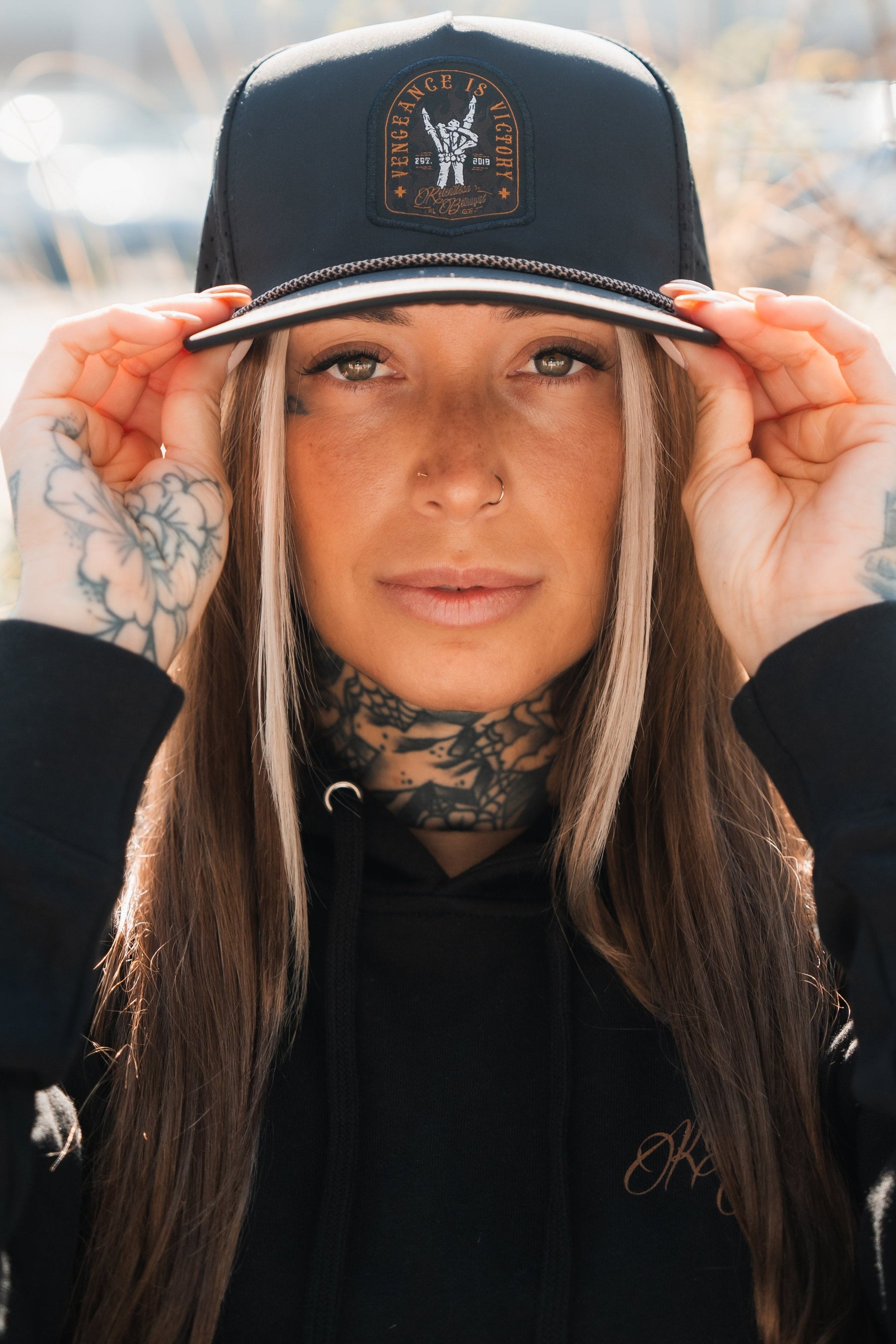 A person with long hair and tattoos on their hands and neck confidently adjusts their black Vengeance Snapback from Relentless Betrayal, emblazoned with Vengeance is Victory, as they look directly at the camera, embodying resilience.