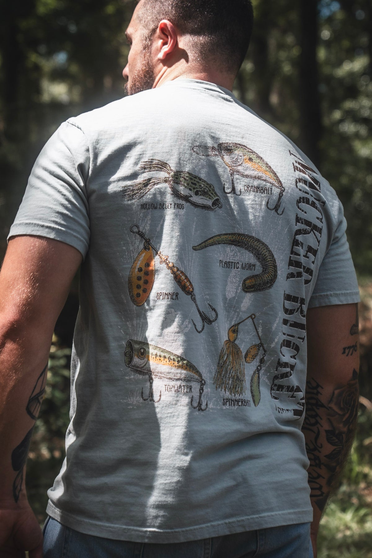 A man stands outdoors in sunlight wearing the Mucker Bucket Bait DNA Premium Tee, crafted from soft ring-spun cotton and featuring detailed illustrations of various fishing lures with their names on the back.