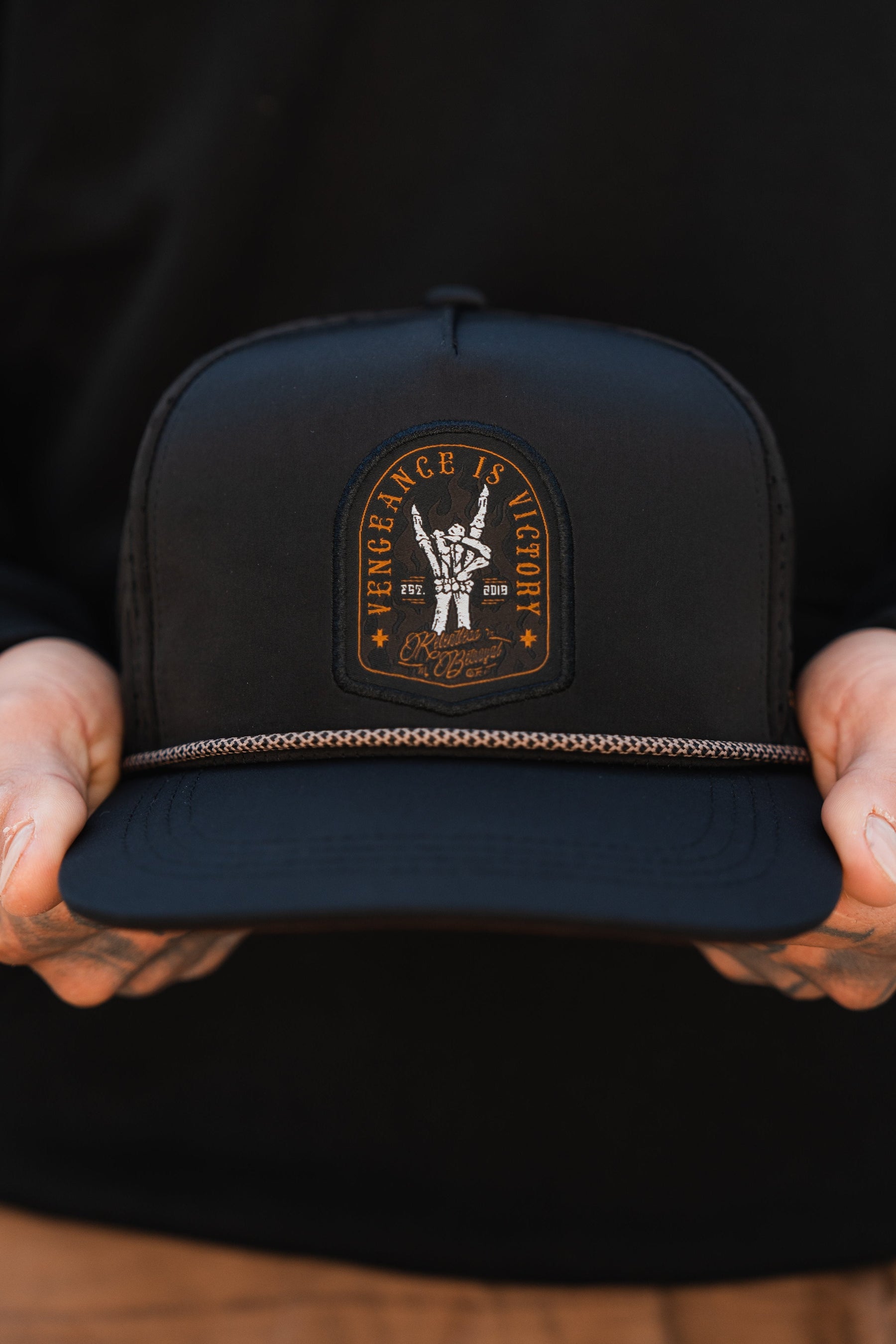A person holds a Vengeance Snapback by Relentless Betrayal, featuring a skeleton hand with a lightning bolt patch symbolizing resilience. The patch states Vengeance is Victory and Dirty Cotton Scoundrels, complemented by a decorative chain above the visor.
