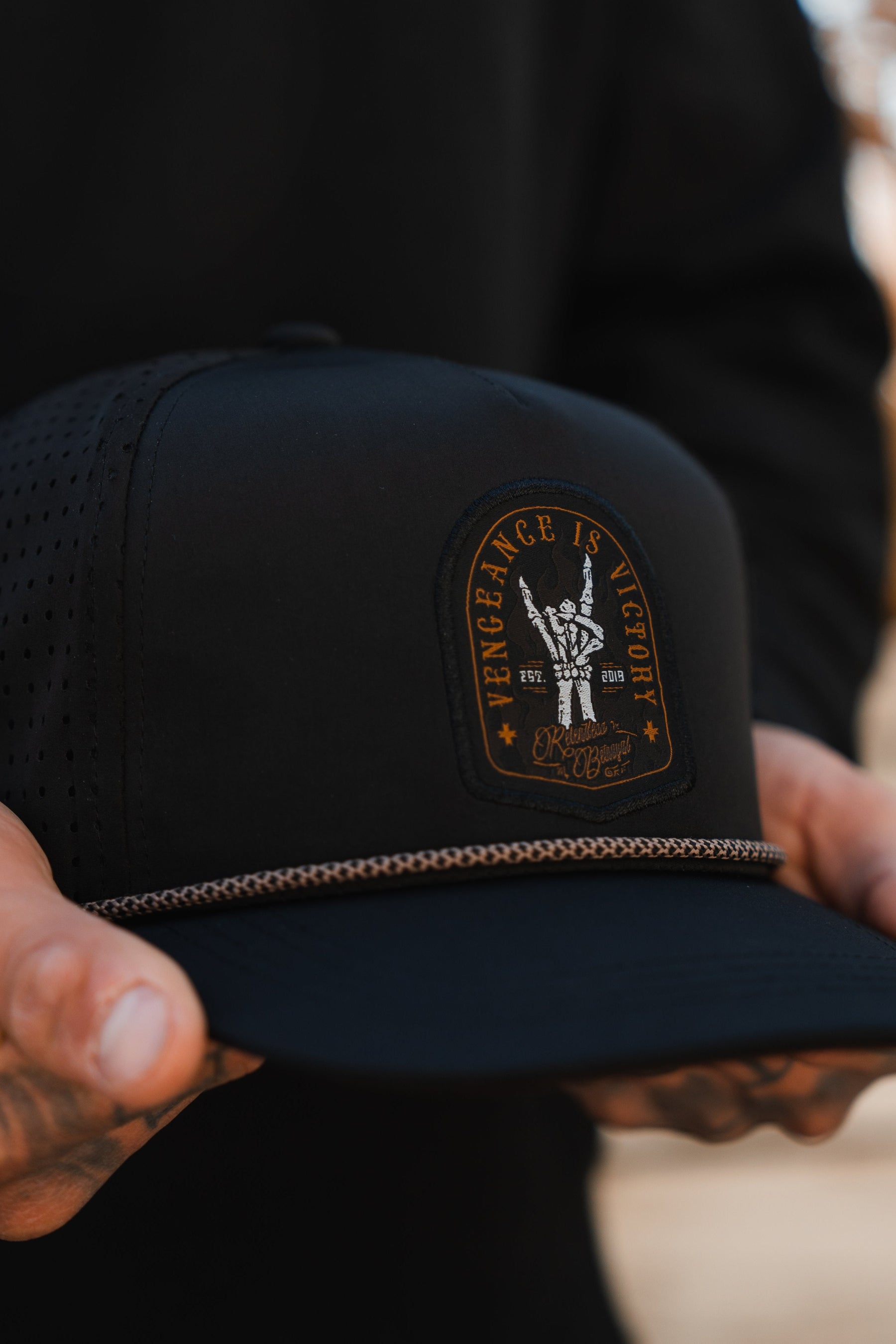 Close-up of a person holding the Vengeance Snapback by Relentless Betrayal, a black cap with a decorative patch featuring a peace-sign hand and Vengeance is Victory text, symbolizing resilience. A thin chain above the brim adds an empowering touch.
