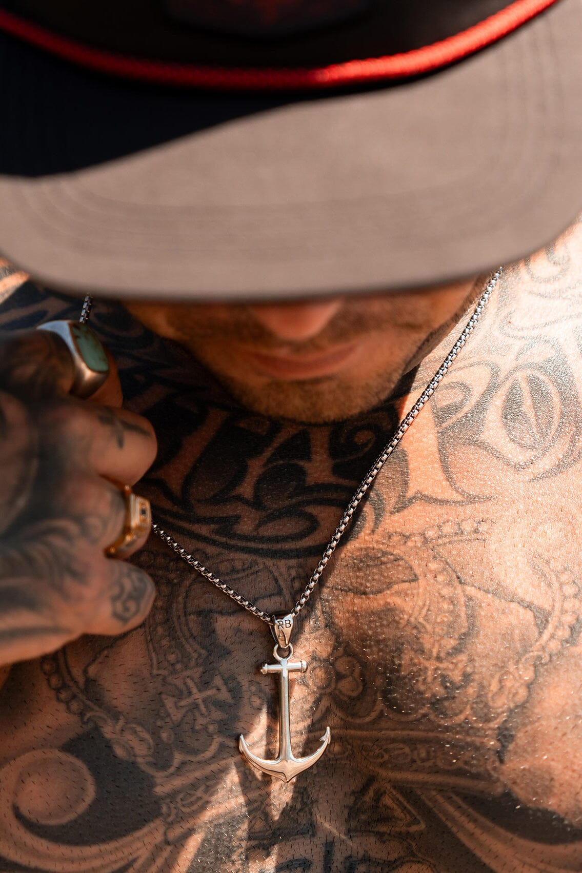 A tattooed person wearing a handcrafted Anchor Drop Necklace by Relentless Betrayal, featuring a pendant on a 26-inch chain, looks down from beneath a wide-brimmed hat. Their ring-adorned hand gently touches the stainless steel necklace.