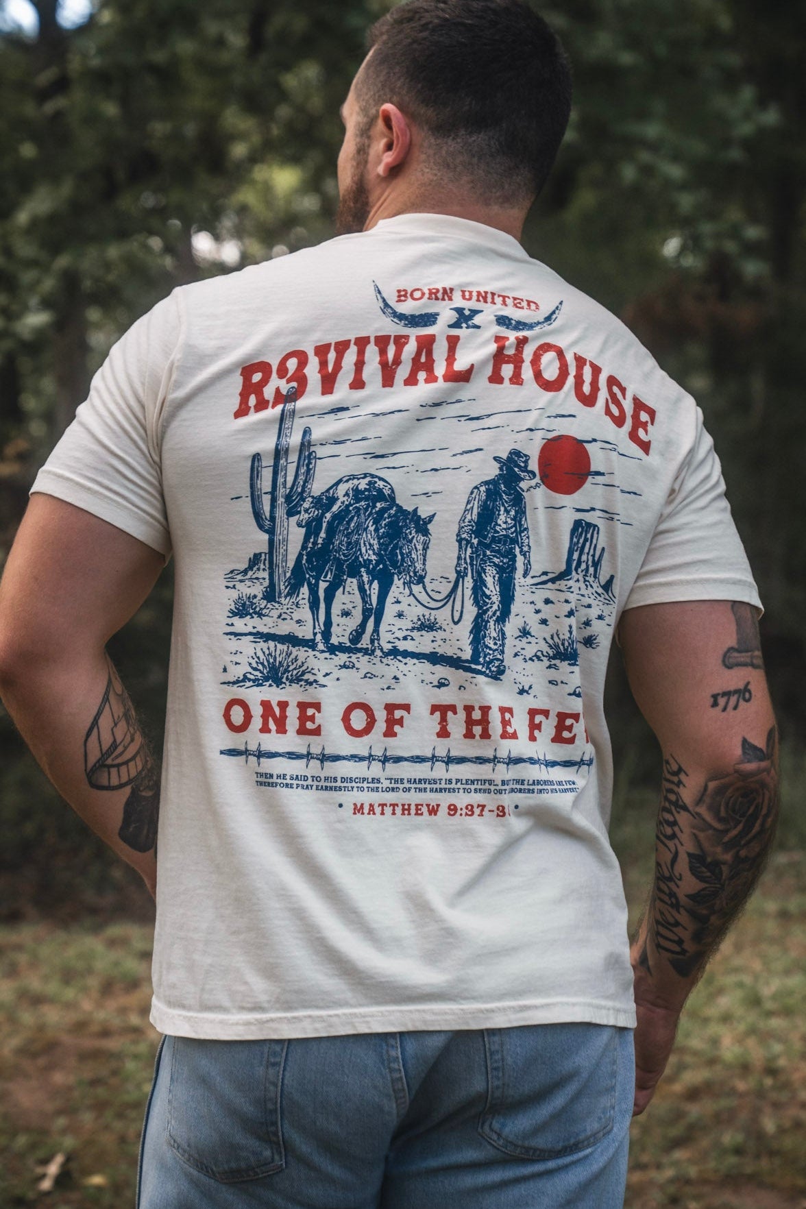 A man stands outdoors with his back to the camera, wearing the Revival House Premium Tee by Born United Private Label—a graphic t-shirt featuring a cowboy and horse, along with the text “Revival House One of the Few” and a Bible verse reference. Inspired by R3VIVAL Ranch, this Christian apparel piece stands out against the backdrop of trees.