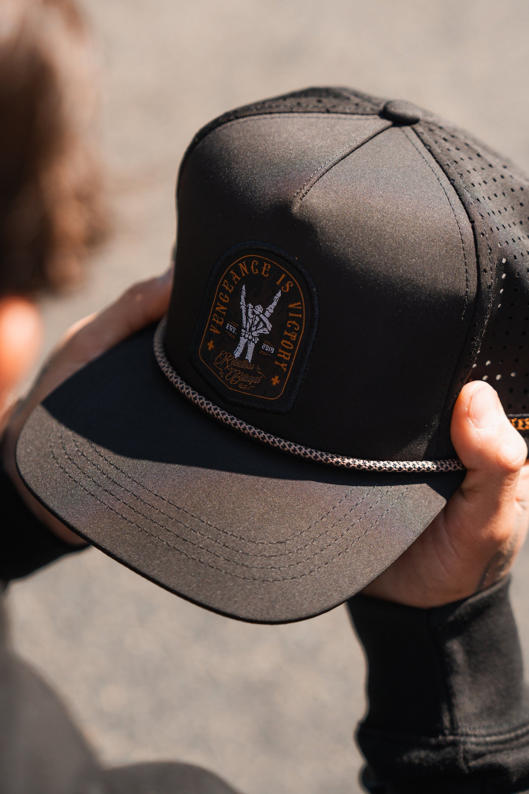 Someone holds a Relentless Betrayals Vengeance Snapback, featuring mesh sides and a gold-lettered Vengeance Is Victory patch with an eagle emblem. A gold chain decorates the brim against a blurred outdoor scene.
