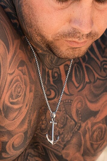 A man with a beard and closed eyes, adorned with rose and script tattoos, wears the Relentless Betrayal handcrafted silver Anchor Drop Necklace, meticulously designed in stainless steel for lasting brilliance.